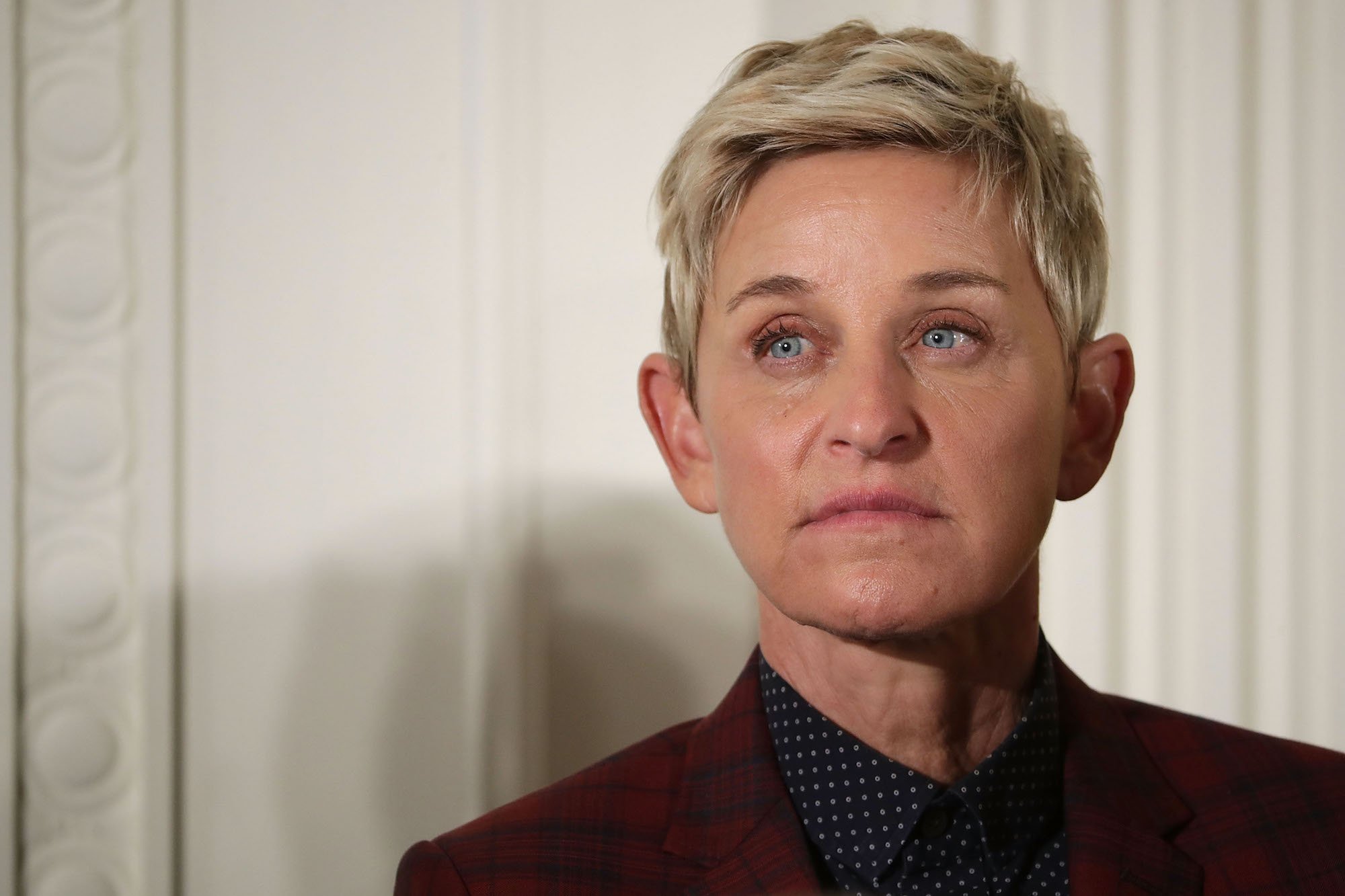 Ellen DeGeneres in front of a white wall not smiling