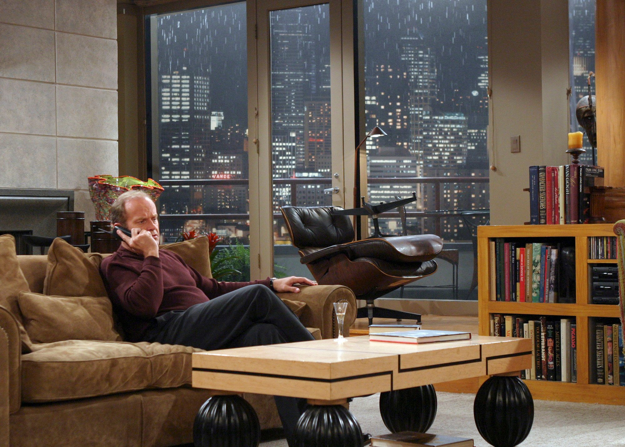 Kelsey Grammer as Dr. Frasier Crane on a couch in a living room, with windows in the background showing rain over a cityscape