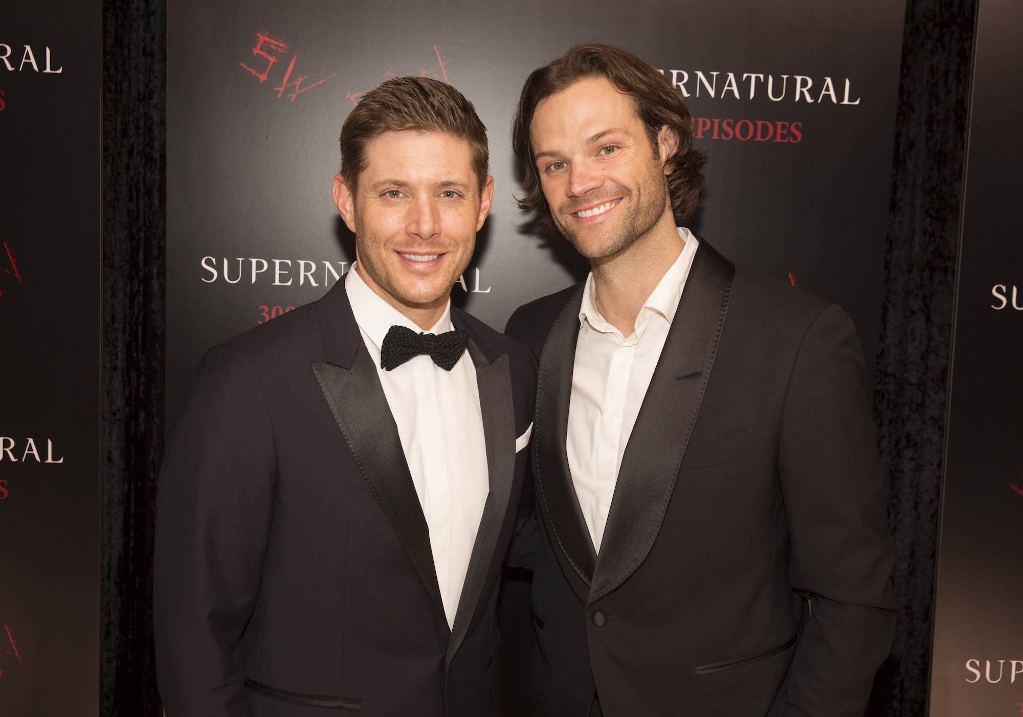 (L-R) Jensen Ackles and Jared Padalecki smiling in front of a black background