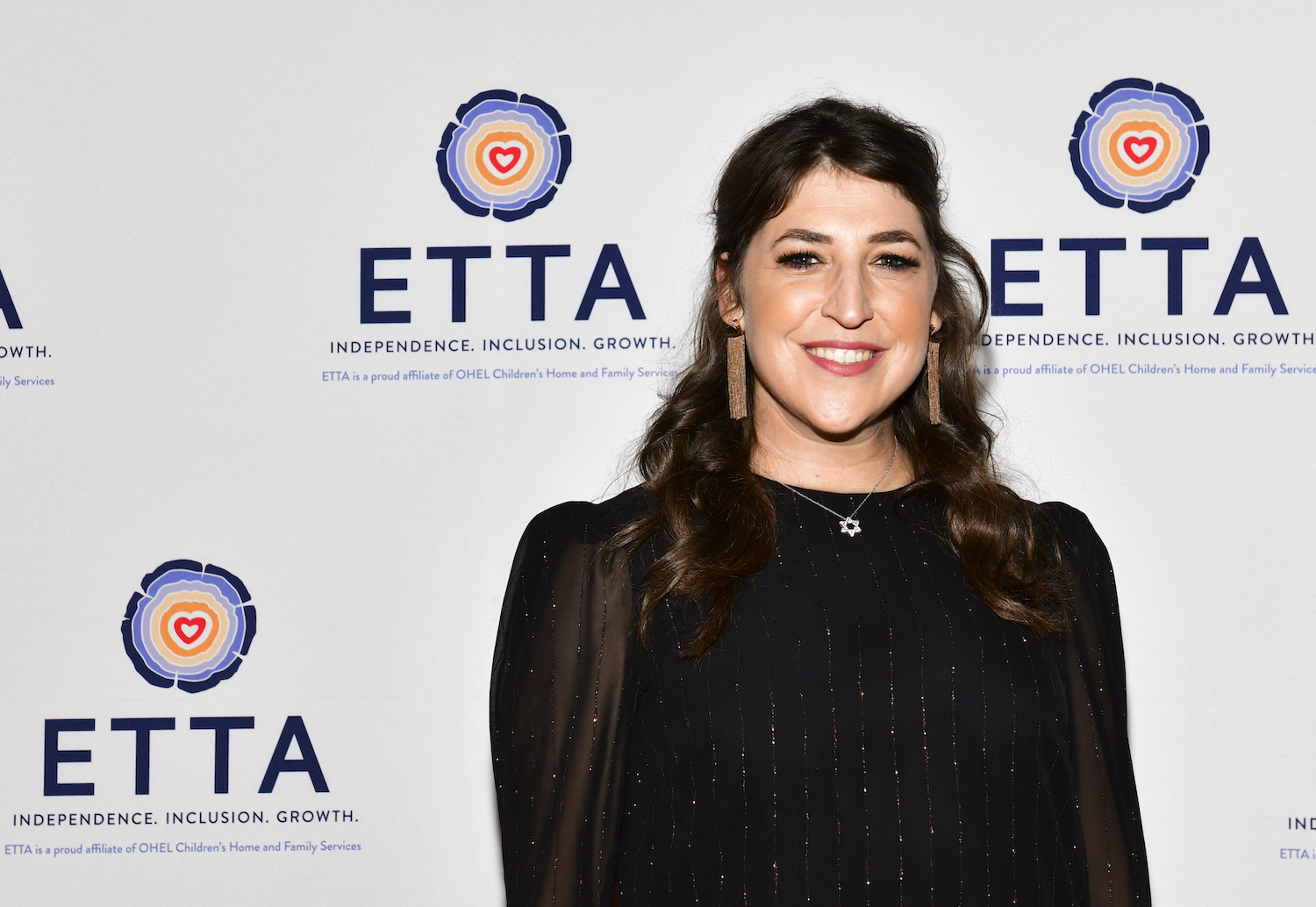 Mayim Bialik smiling in front of a white background