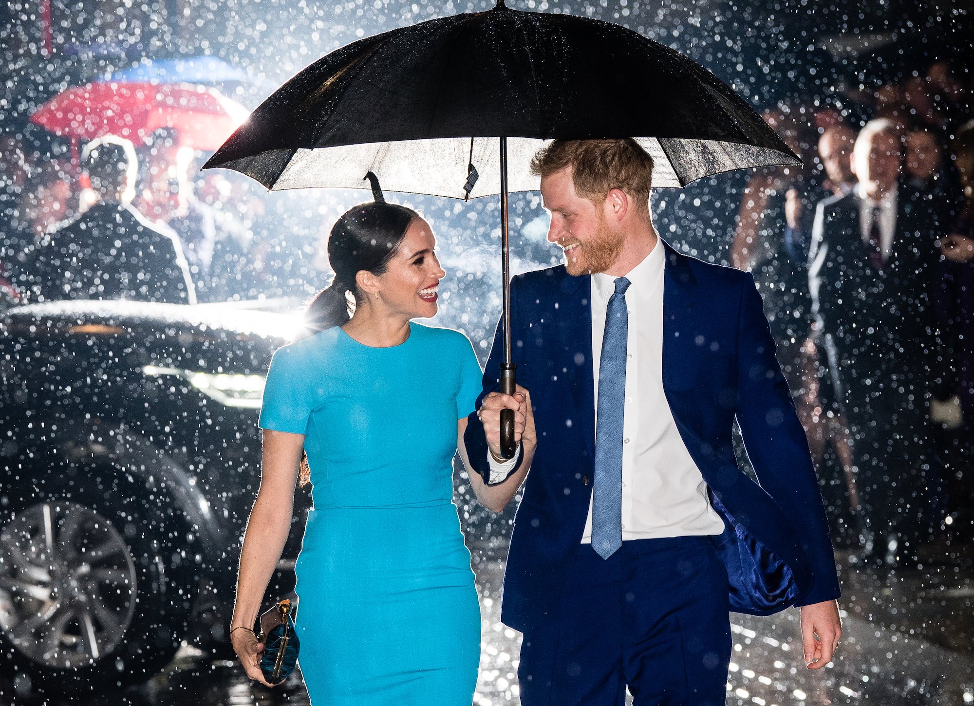 (L-R) Meghan Markle and Prince Harry smiling at each other under and umbrella in the rain