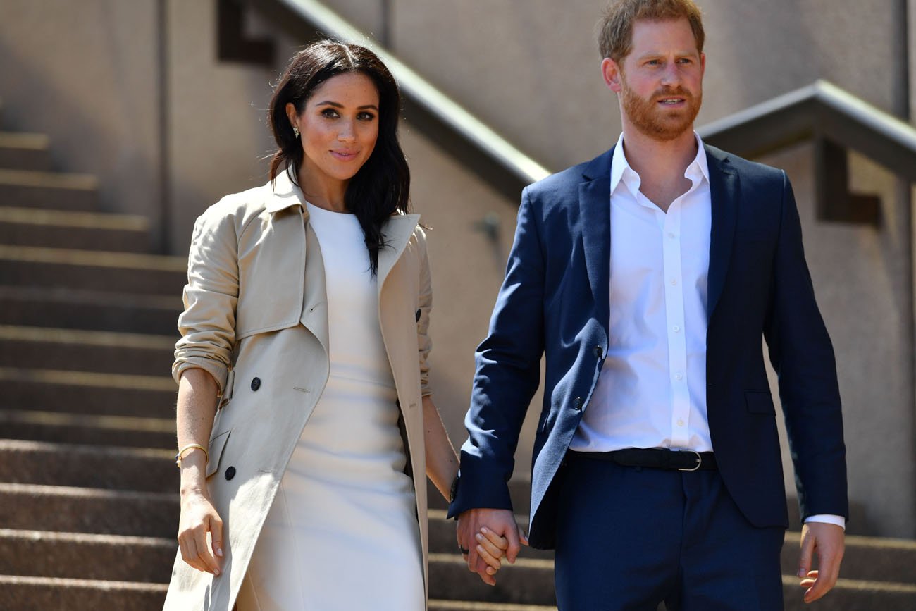 Meghan Markle wears a white dress and tan overcoat while holding hands with Prince Harry dressed in a suit as they walk down stairs