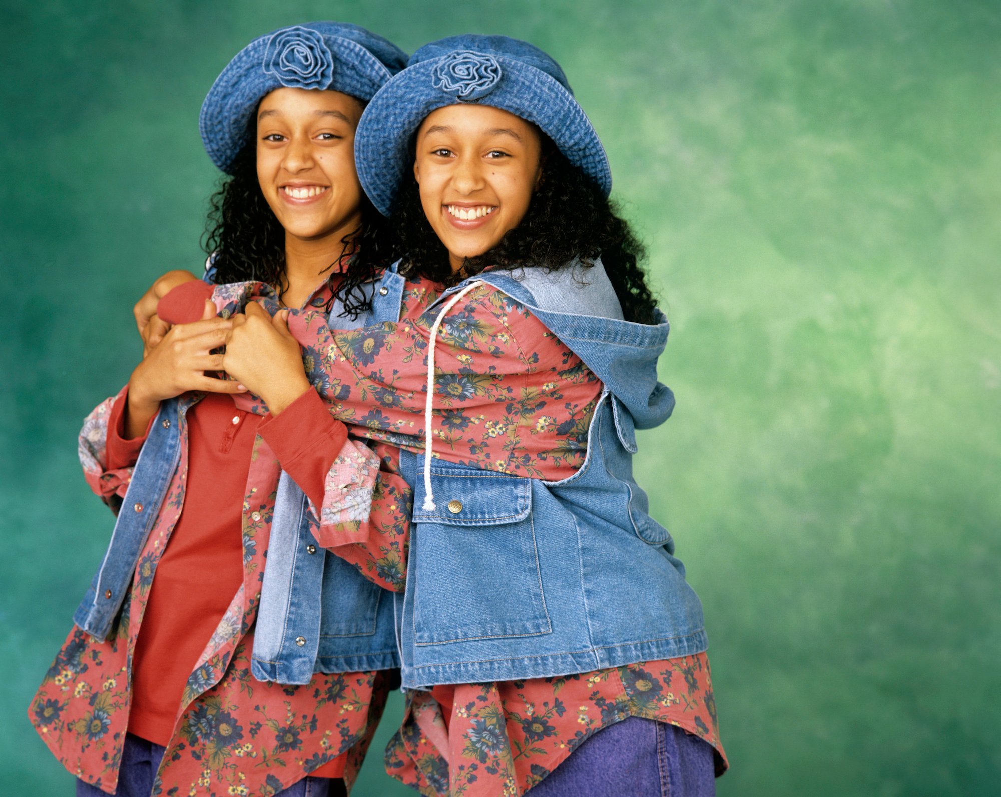 (L-R) Tia Mowry and Tamera Mowry smiling in front of a green background