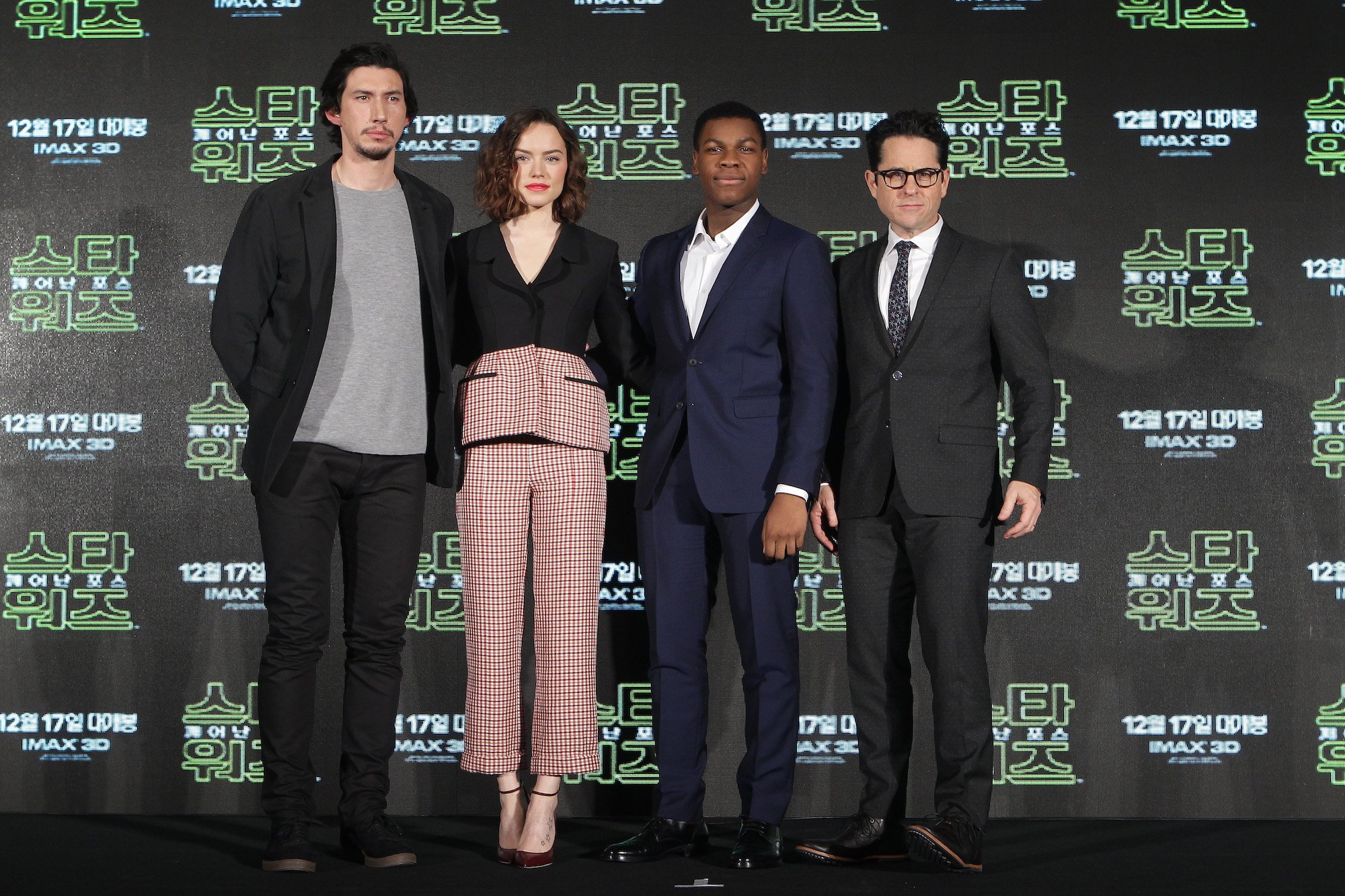 (L to R) Adam Driver, Daisy Ridley, John Boyega and director J.J. Abrams smiling in front of a patterned background