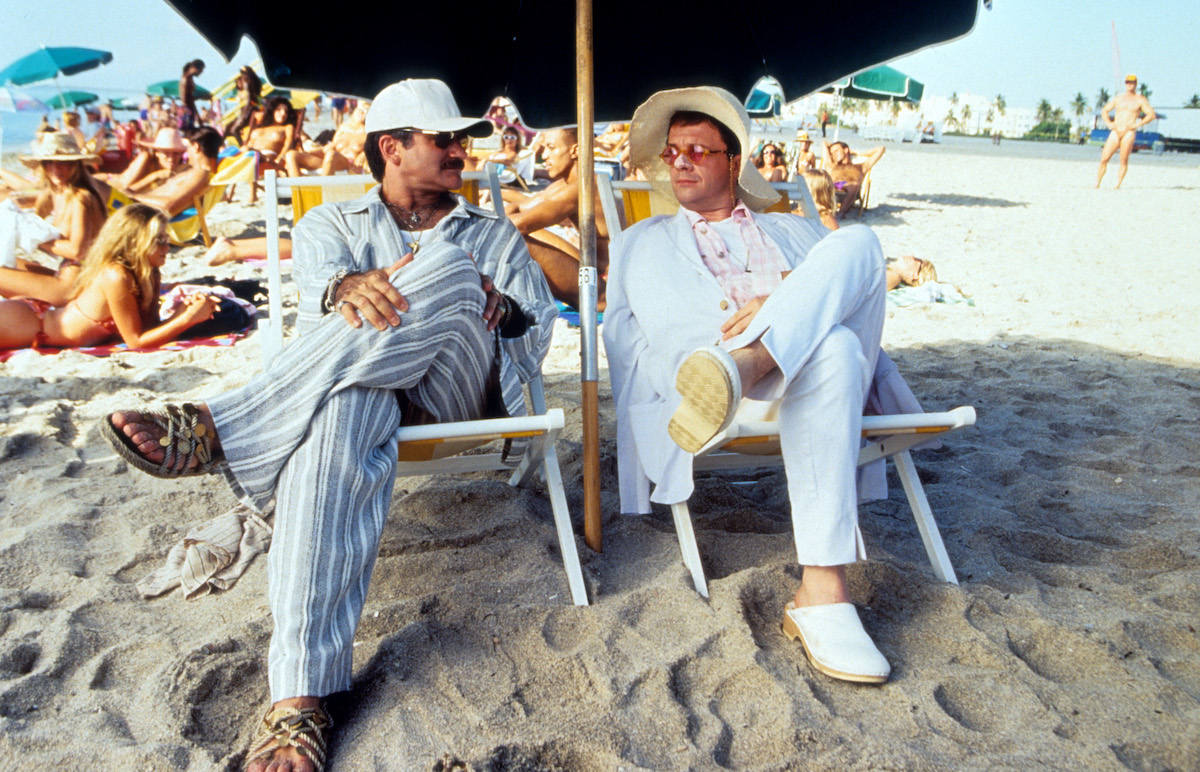 Robin Williams and Nathan Lane sitting under an umbrella on the sand at the beach in a scene from the film 'The Birdcage', 1996