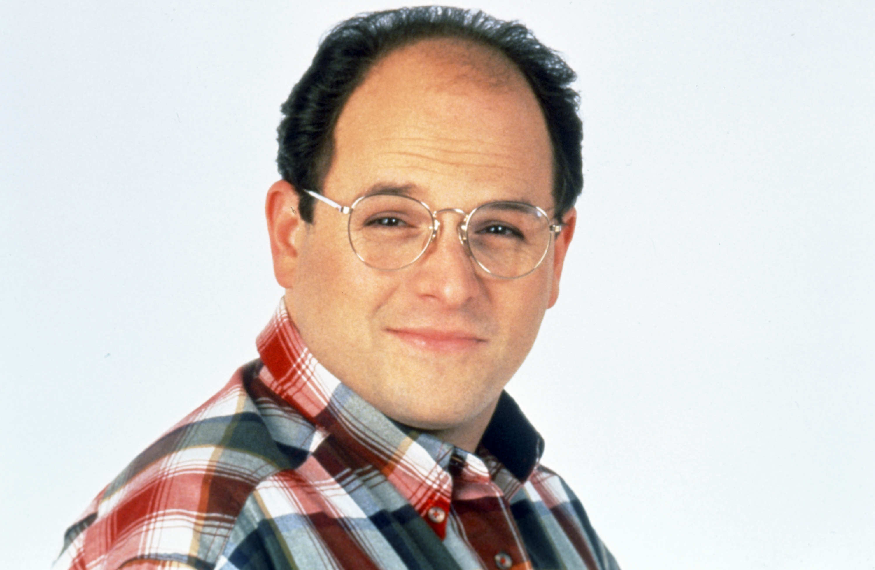 Jason Alexander wearing glasses and a collared shirt while portraying George Costanza