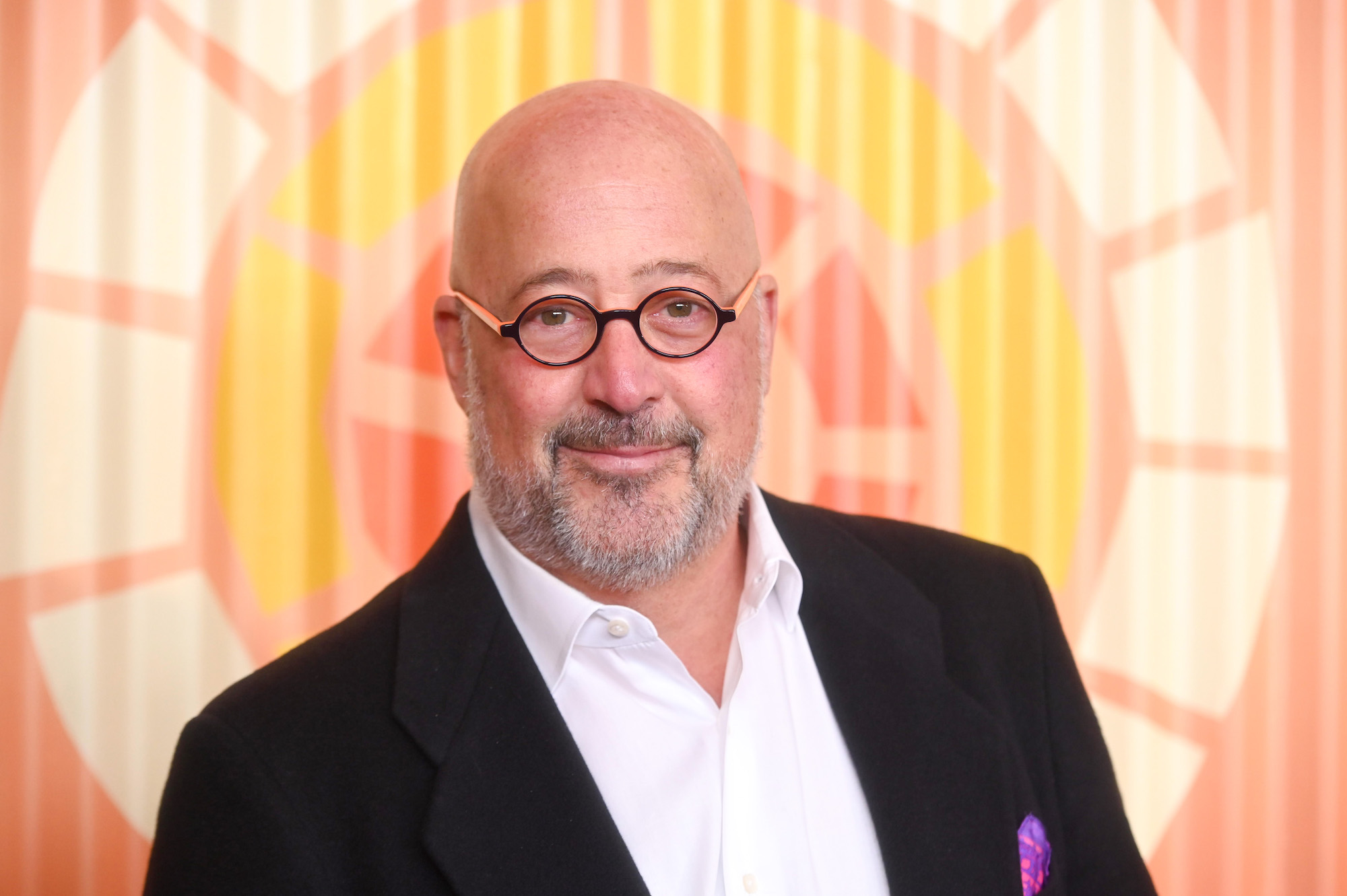 Andrew Zimmern smiling in front of an orange background
