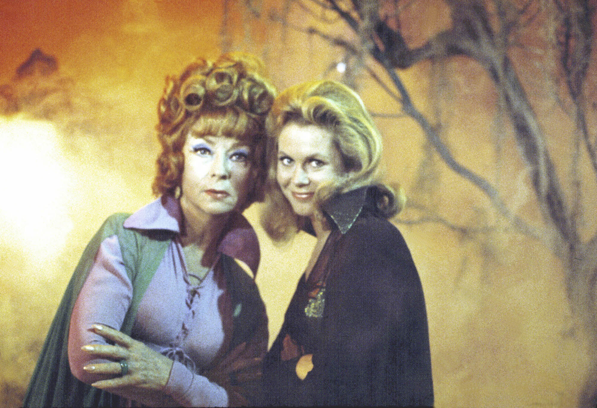 (L-R) Agnes Moorehead as Endora and Elizabeth Montgomery as Samantha Stephens smiling, wearing capes, in front of an orange-lit forest
