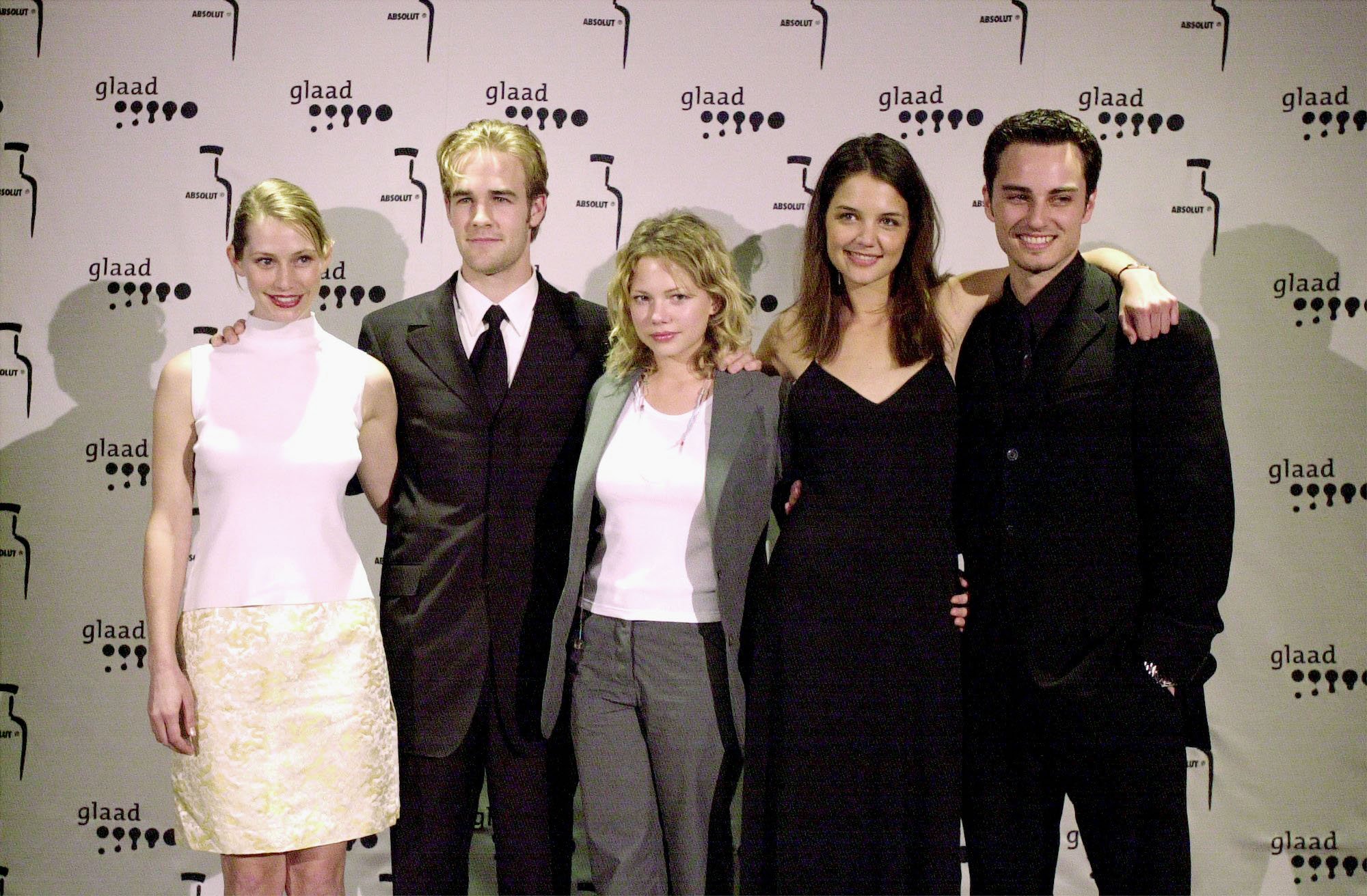 (L-R) Meredith Monroe, James Van der Beek, Michelle Williams, Katie Holmes, and Kerr Smith smiling in front of a white backgrounf