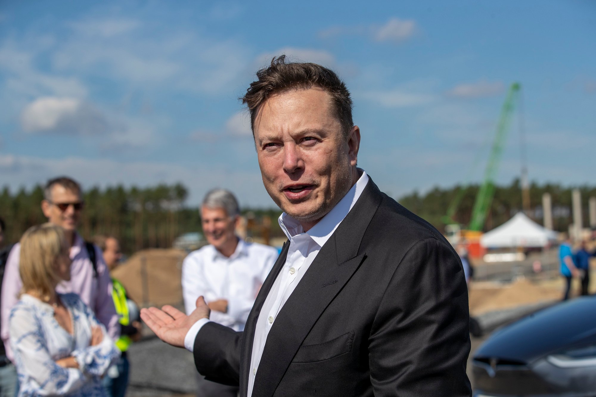 Elon Musk smiling, gesturing with his arm extendedbehind him
