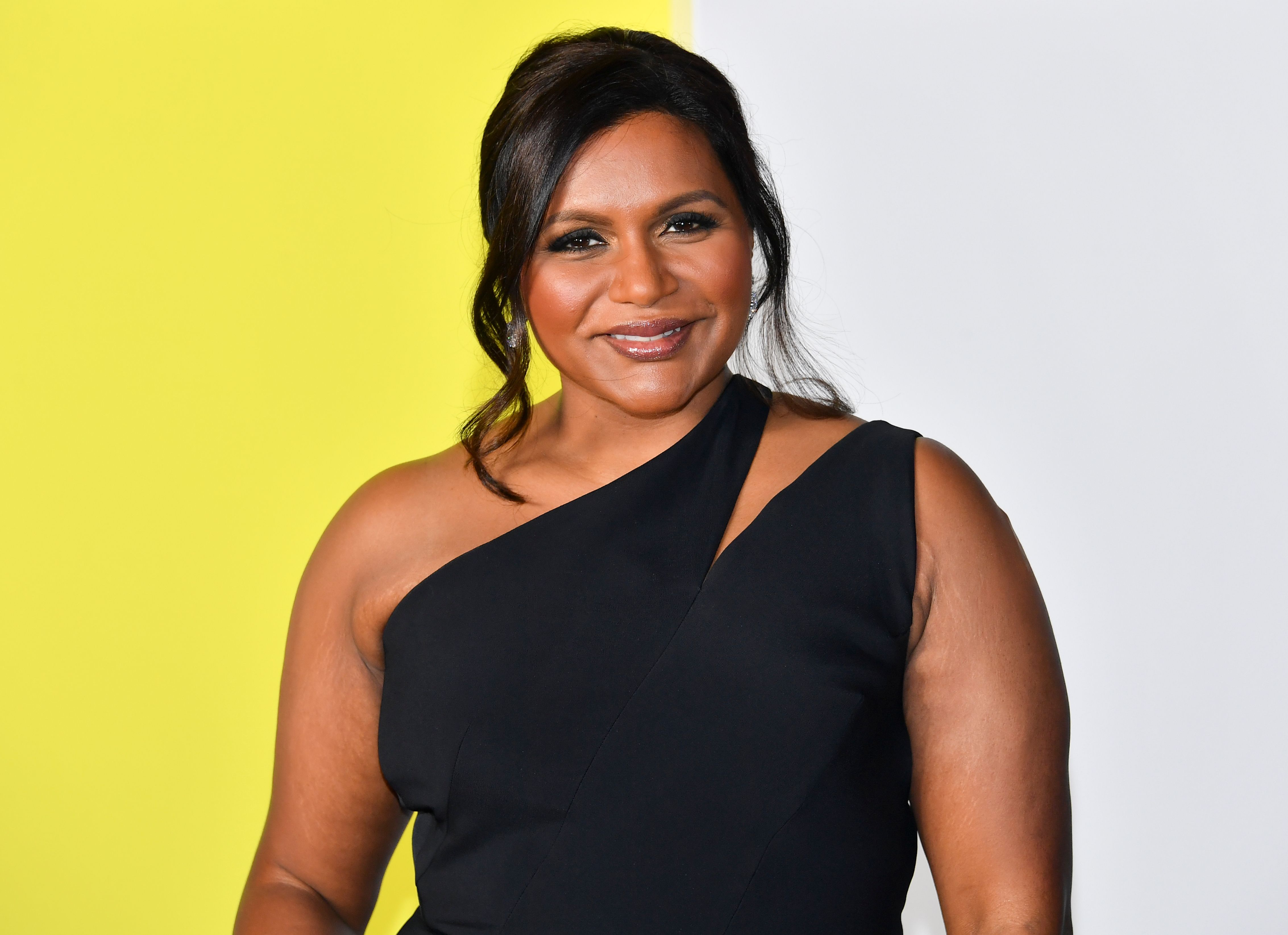 Mindy Kaling poses in a black dress in front of a yellow and gray background. Mindy Kaling husband does not exist so who is the father of Mindy Kaling baby?