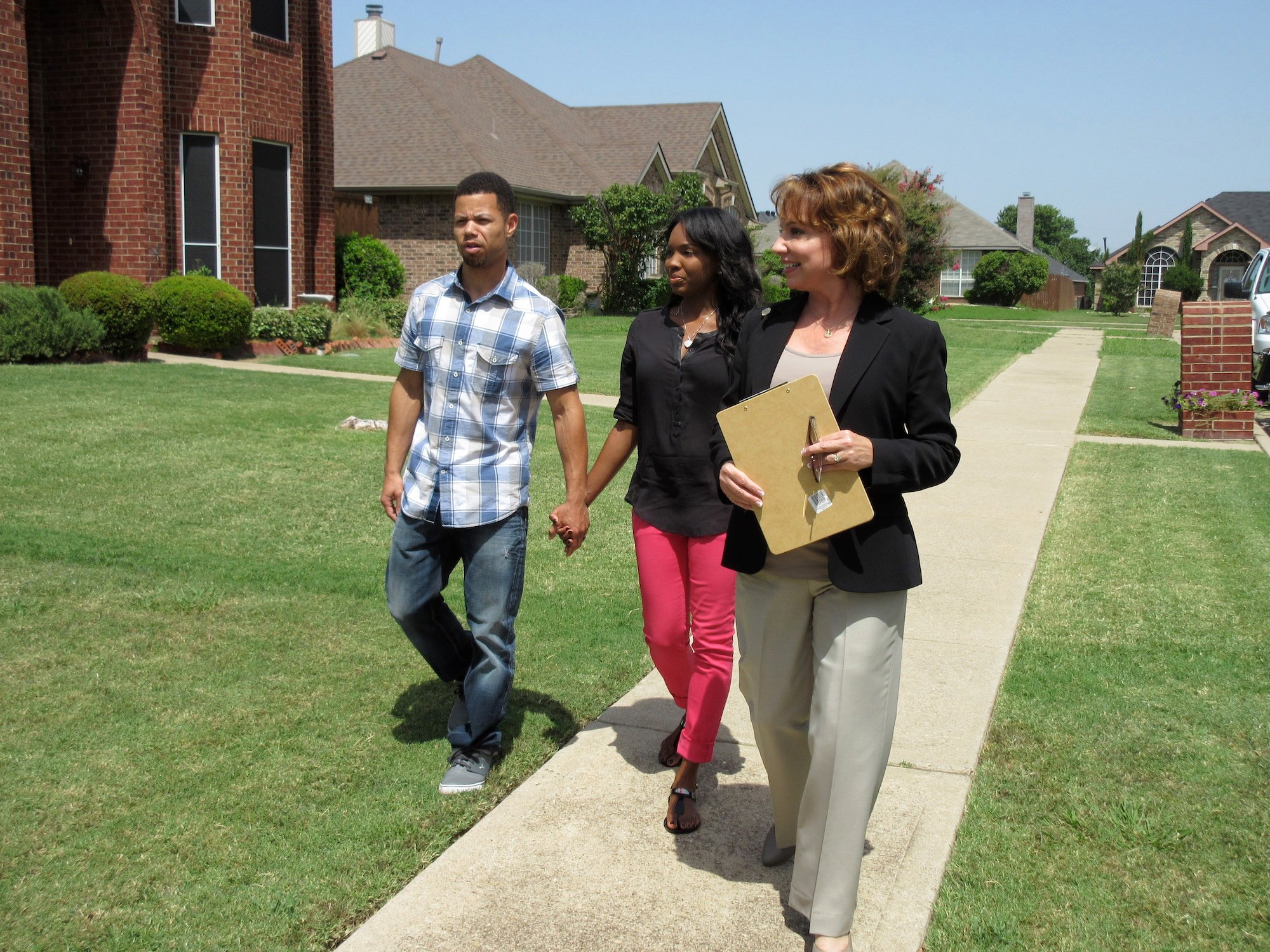 A young couple holding hands walking down a sidewalk with a realtor on 'House Hunters'