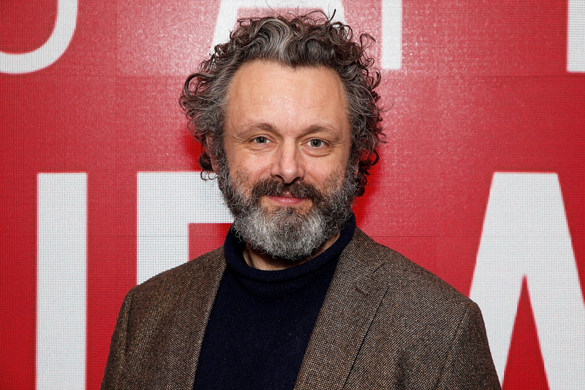 Michael Sheen smiling in front of a red background