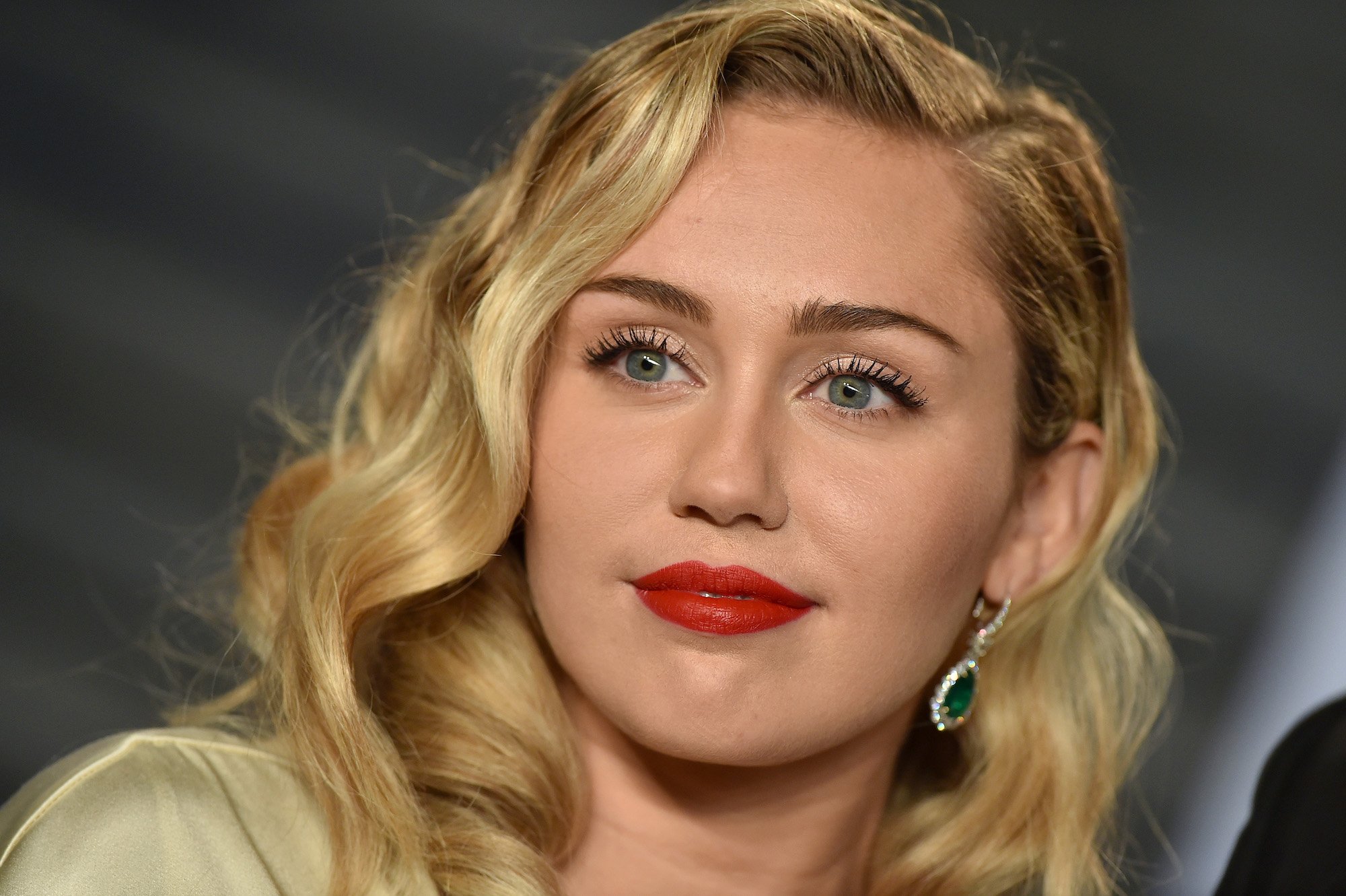 Miley Cyrus slightly smiling, wearing red lipstick, in front of a blurred background