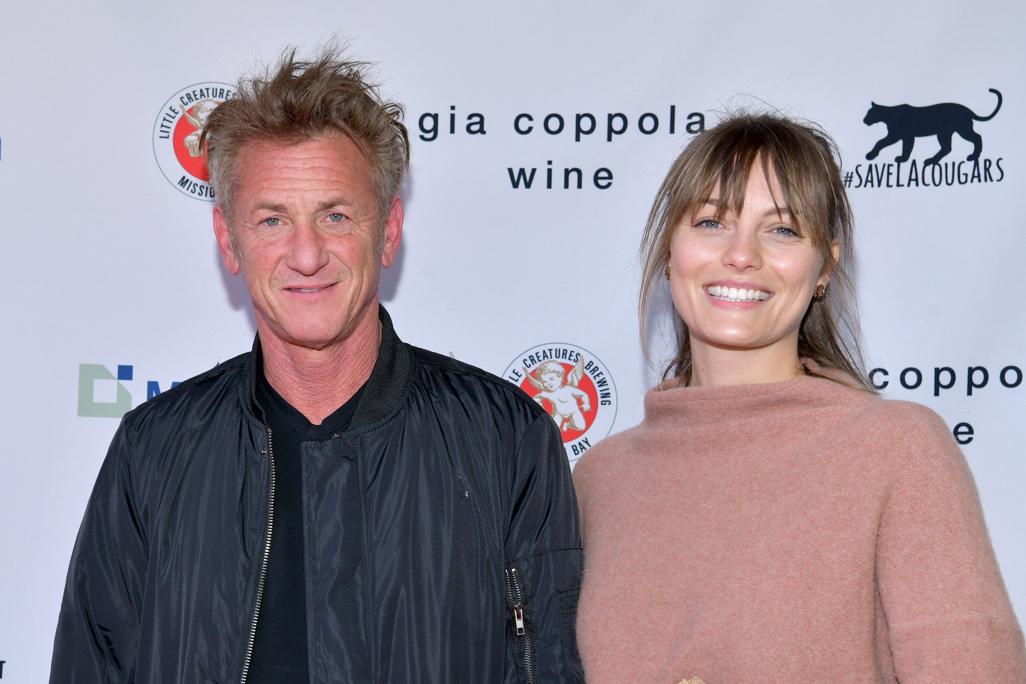 (L-R) Sean Penn and Leila George smiling in front of a white background with black and red logos