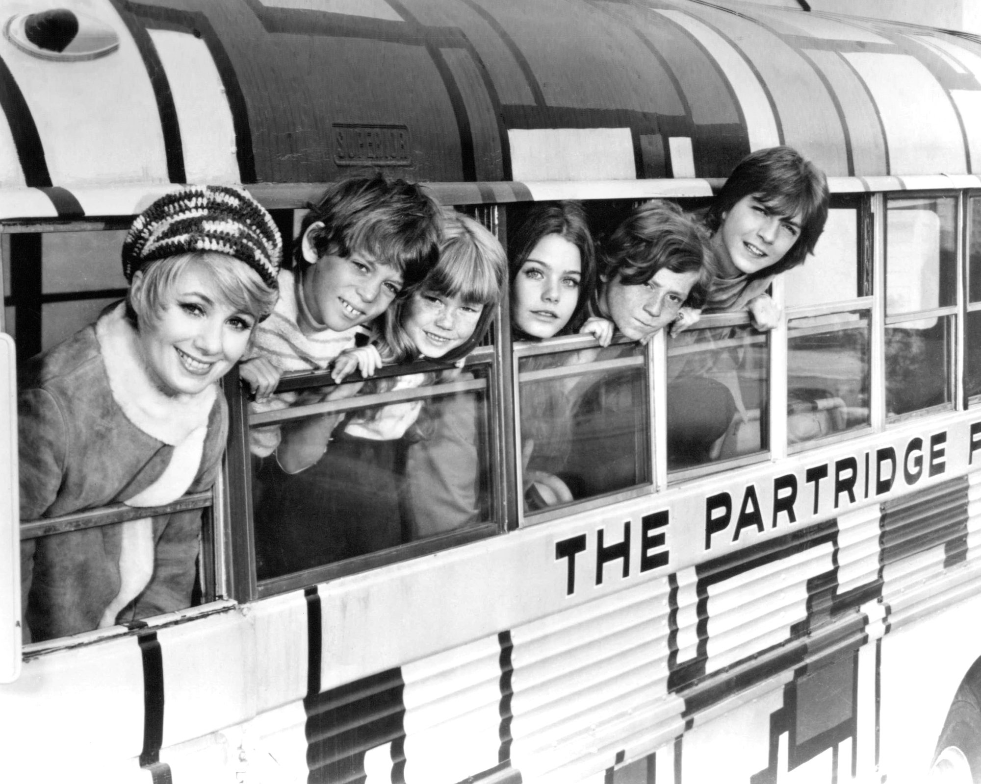 (L-R) Shirley Jones, Jeremy Gelbwaks, Suzanne Crough, Susan Dey, Danny Bonaduce and David Cassidy leaning out of schoolbus windows, in black and white