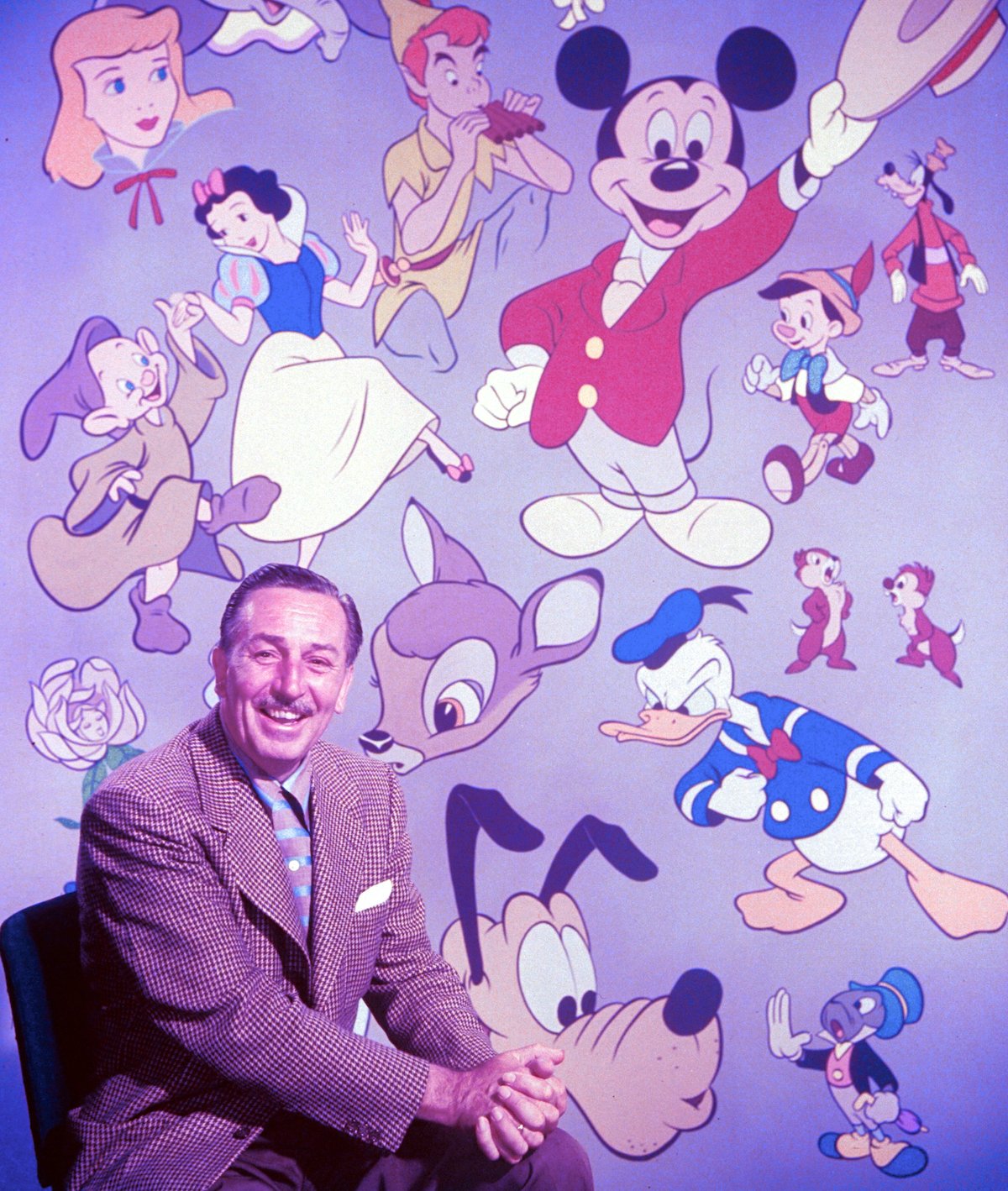 Walt Disney sitting in front of backdrop filled with Disney creations