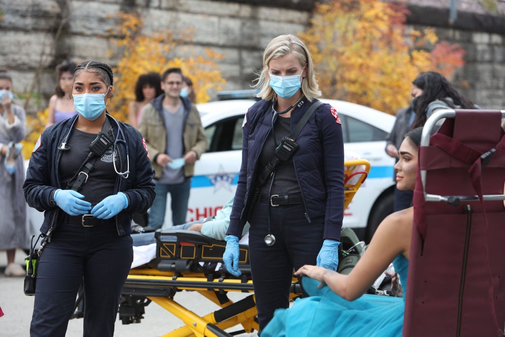 (L-R) Adriyan Rae as Gianna Mackey, Kara Killmer as Sylvie Brett wearing masks, standing outside in front of a small crowd
