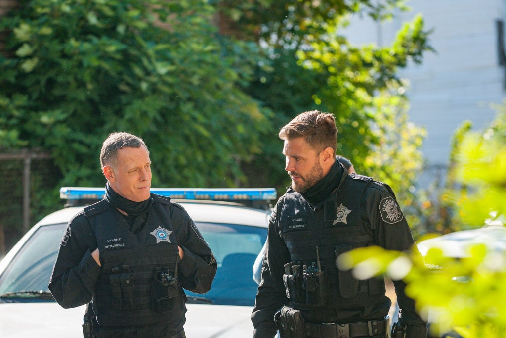 (L-R) Jason Beghe as Hank Voight, Patrick John Flueger as Adam Ruzek turned toward each other, in front of a cop car and tree