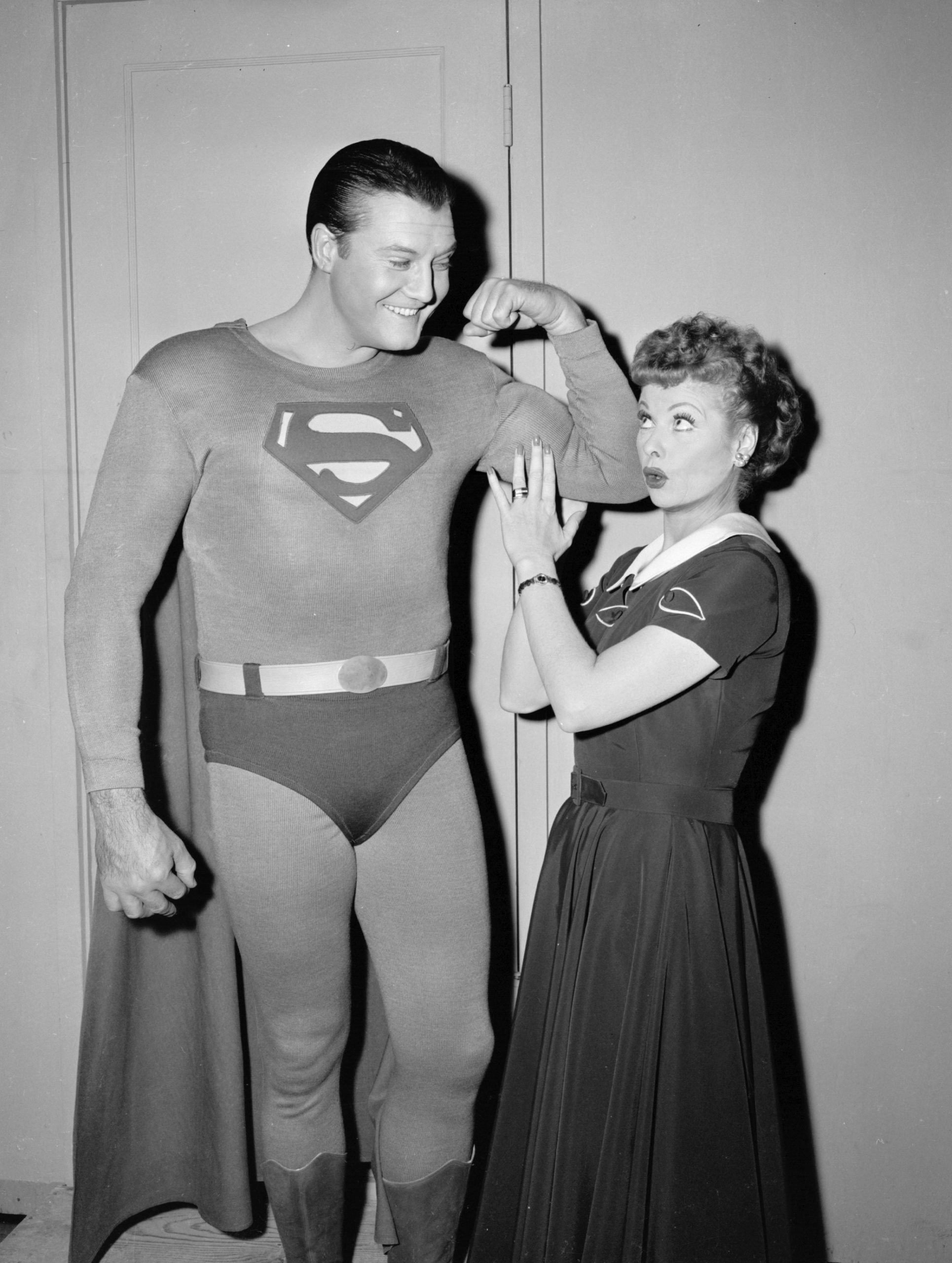 George Reeves flexes his bicep in a Superman costume while Lucille Ball touches his arm on the set of 'I Love Lucy'