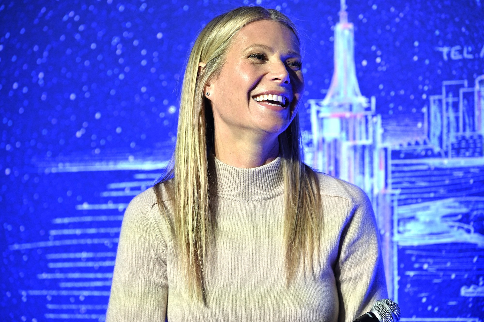 Gwyneth Paltrow laughing, looking to the right, sitting in front of a blue background