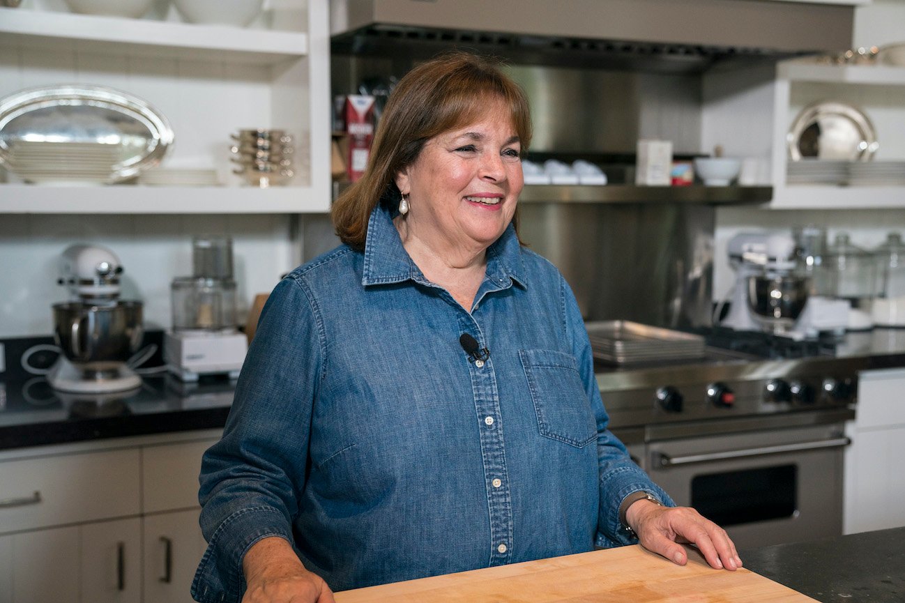 Ina Garten smiles standing in front of a butcher block wearing a denim shirt