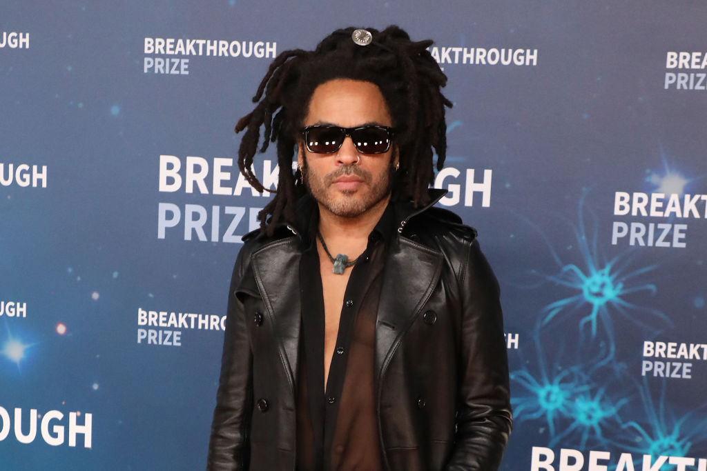 Lenny Kravitz standing in front of a blue background, wearing sunglasses