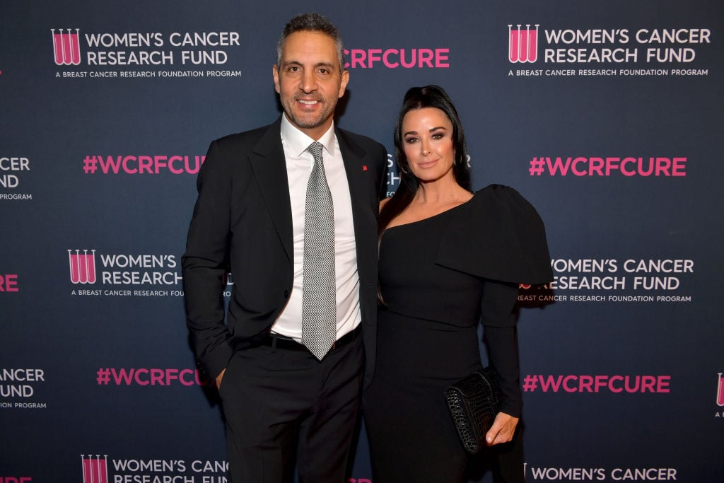 (L-R) Mauricio Umansky and Kyle Richards smiling in front of a dark background with pink logos