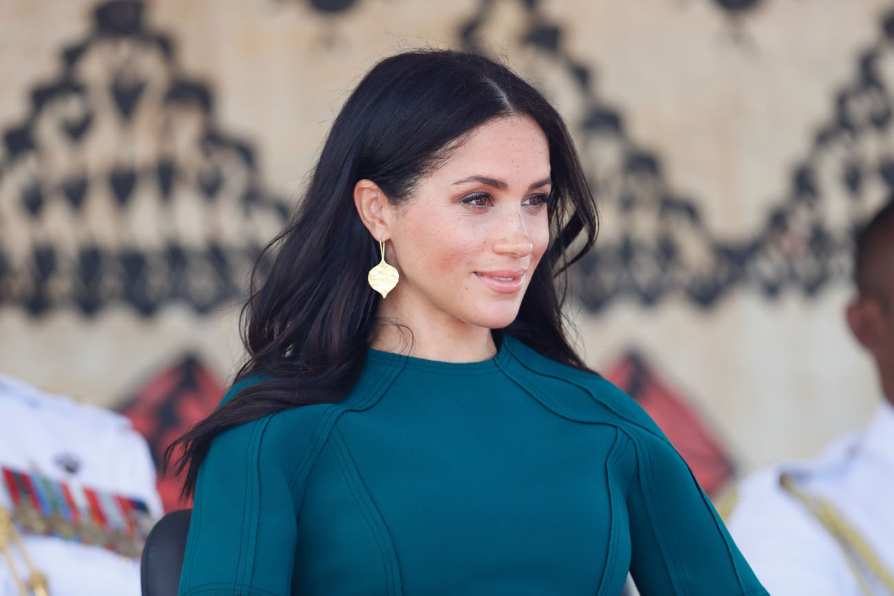 Meghan Markle looking on in a teal dress with gold earrings