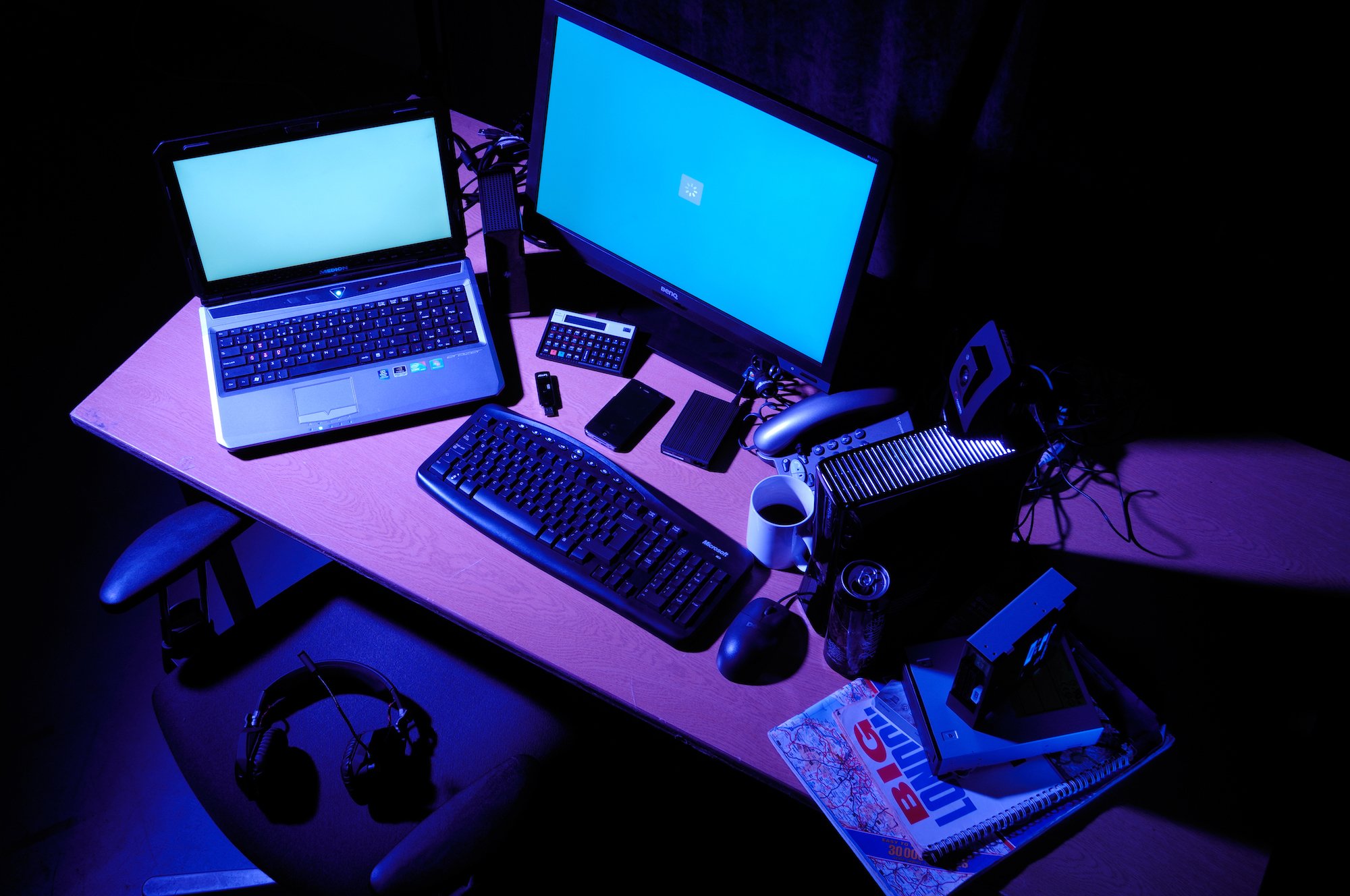 Laptop and desktop computer sitting on a desk with a coffee cup, magazines, pens, and a keyboard