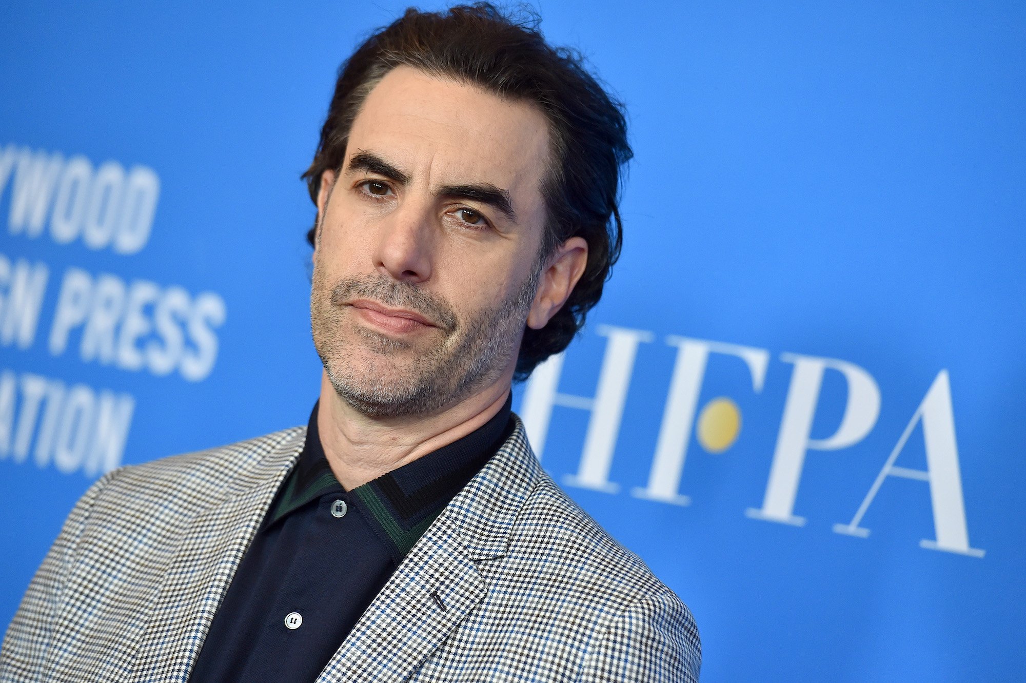 Sacha Baron Cohen smiling slightly in front of a blue background