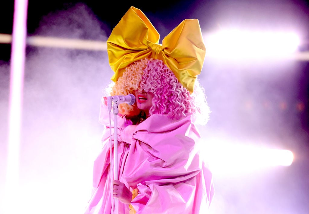 Sia wearing a curly pink wig and yellow bow performing on stage