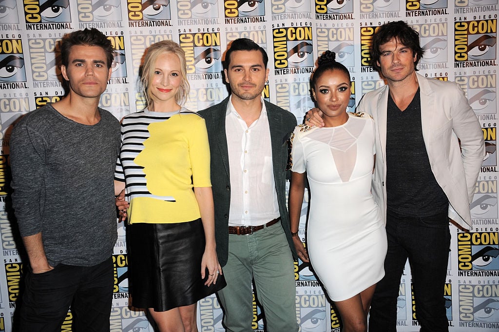 (L-R) Paul Wesley, Candice Accola, Michael Malarkey, Kat Graham and Ian Somerhalder smiling in front of a repeating background with the Comic Con logo