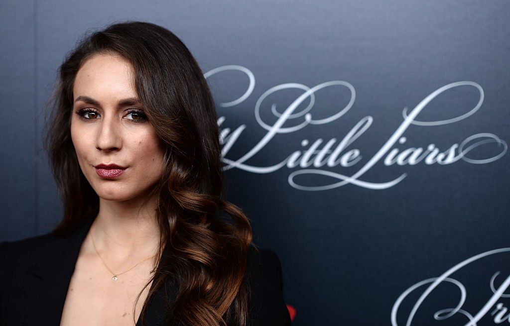 Troian Bellisario slightly smiling in front of a black background with Pretty Little Liars logo