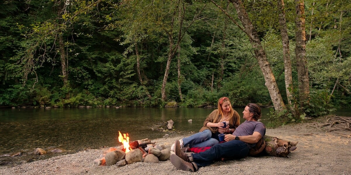 Mel and Jack sitting on on a riverbank in front of a fire in 'Virgin River' Season 5