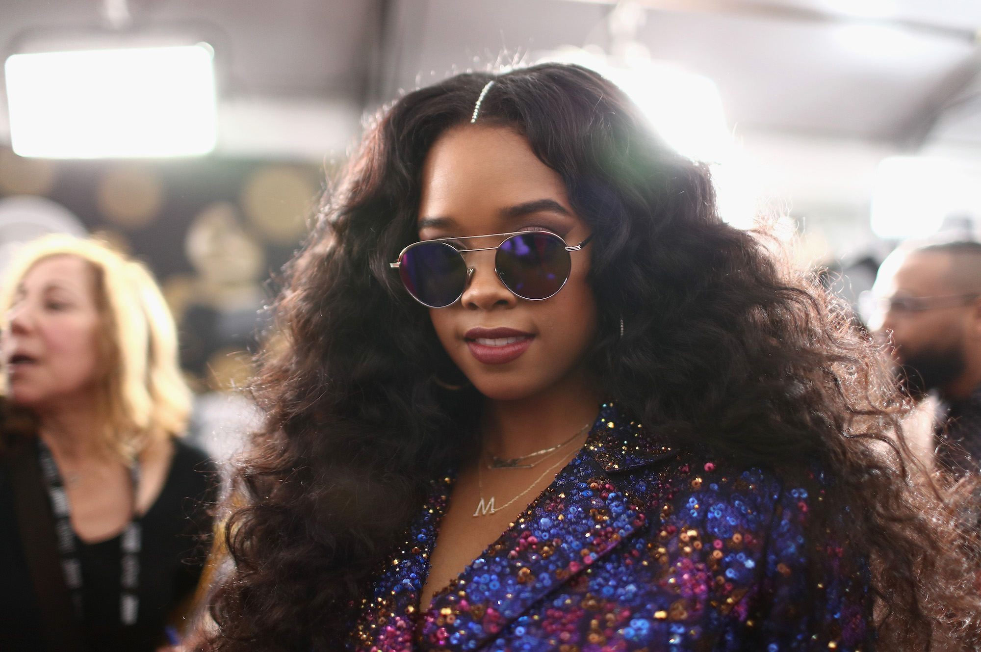 H.E.R. smiling, wearing sunglasses, in front of a blurred crowd