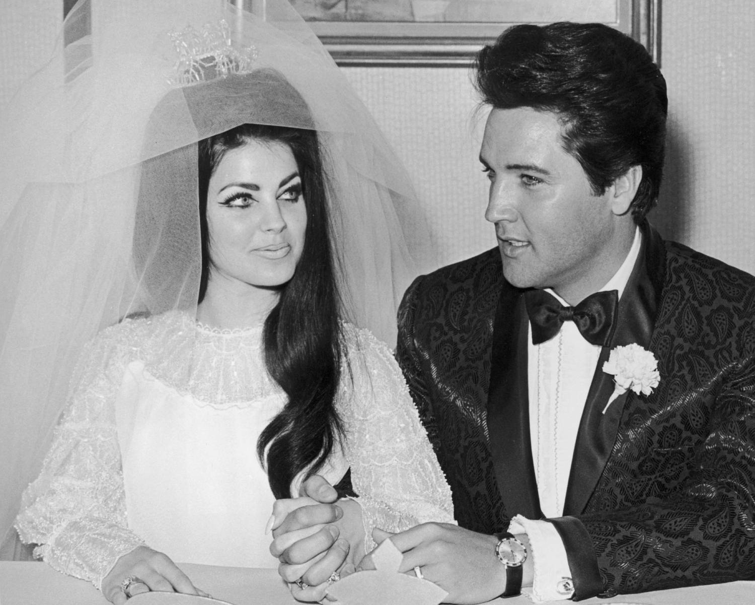 American rock n' roll singer and actor Elvis Presley (1935 - 1977) sits and holds hands with his bride Priscilla Presley on their wedding day, Las Vegas, Nevada. She wears her wedding gown and veil. He wears a carnation in his tuxedo lapel.