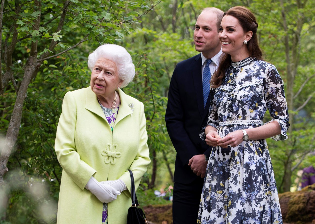 Queen Elizabeth II stands next to Prince William and Kate Middleton with green foliage in the background