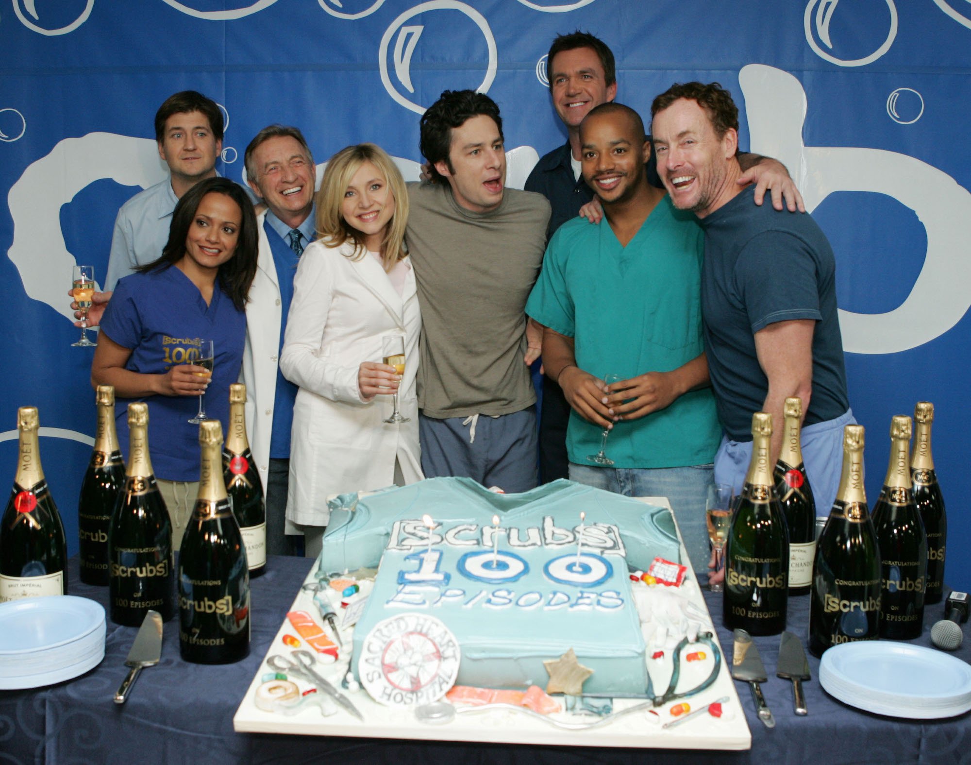 (L-R) Judy Reyes, producer Bill Lawrence, Ken Jenkins, Sarah Chalke, Zach Braff, Neil Flynn, Donald Faison and John C. McGinley smiling in front of a cake celebrating the 100th episode
