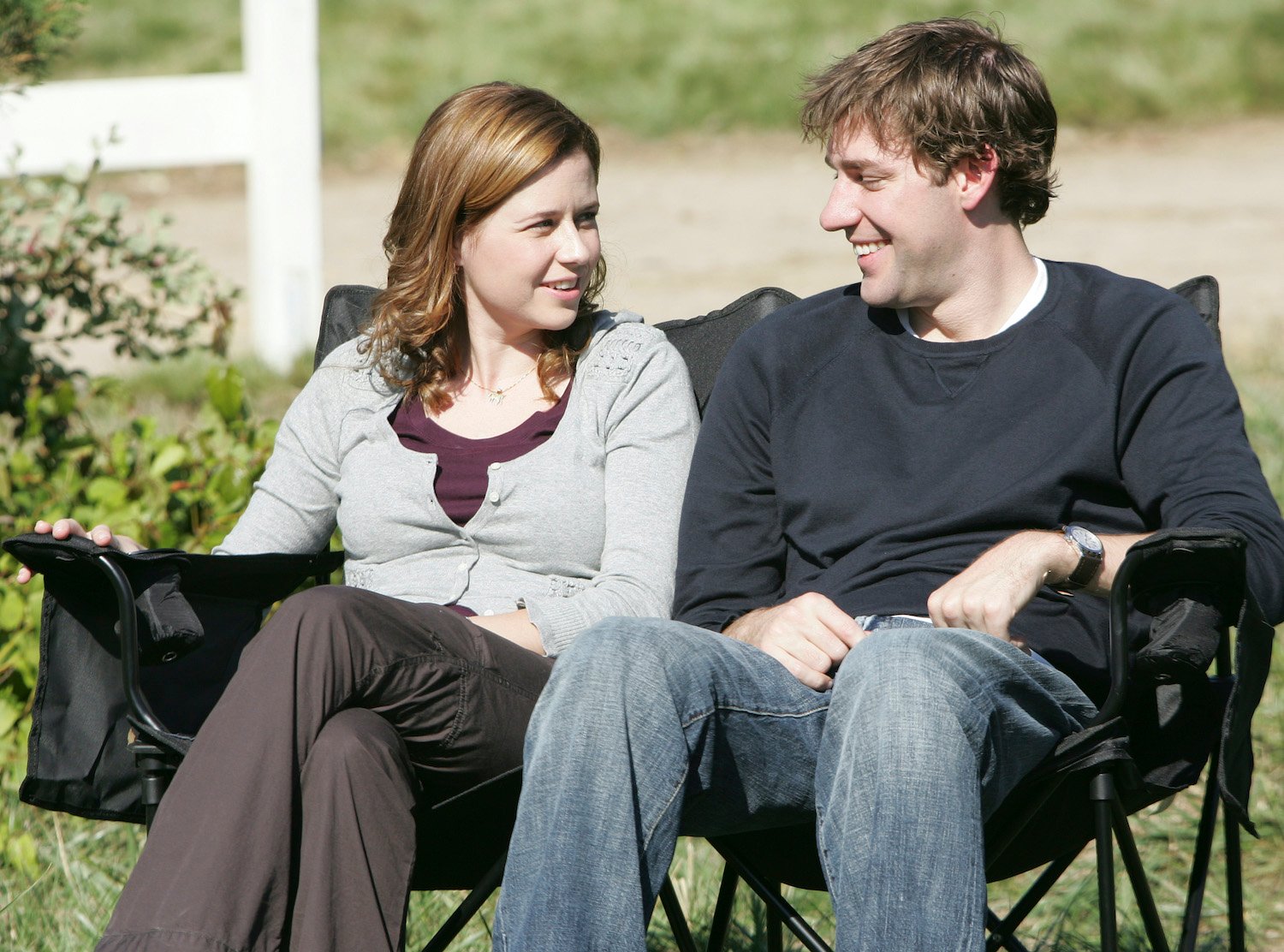 The Office stars Jenna Fischer as Pam Beesly and John Krasinski as Jim Halpert sit in chairs and smile at each other.