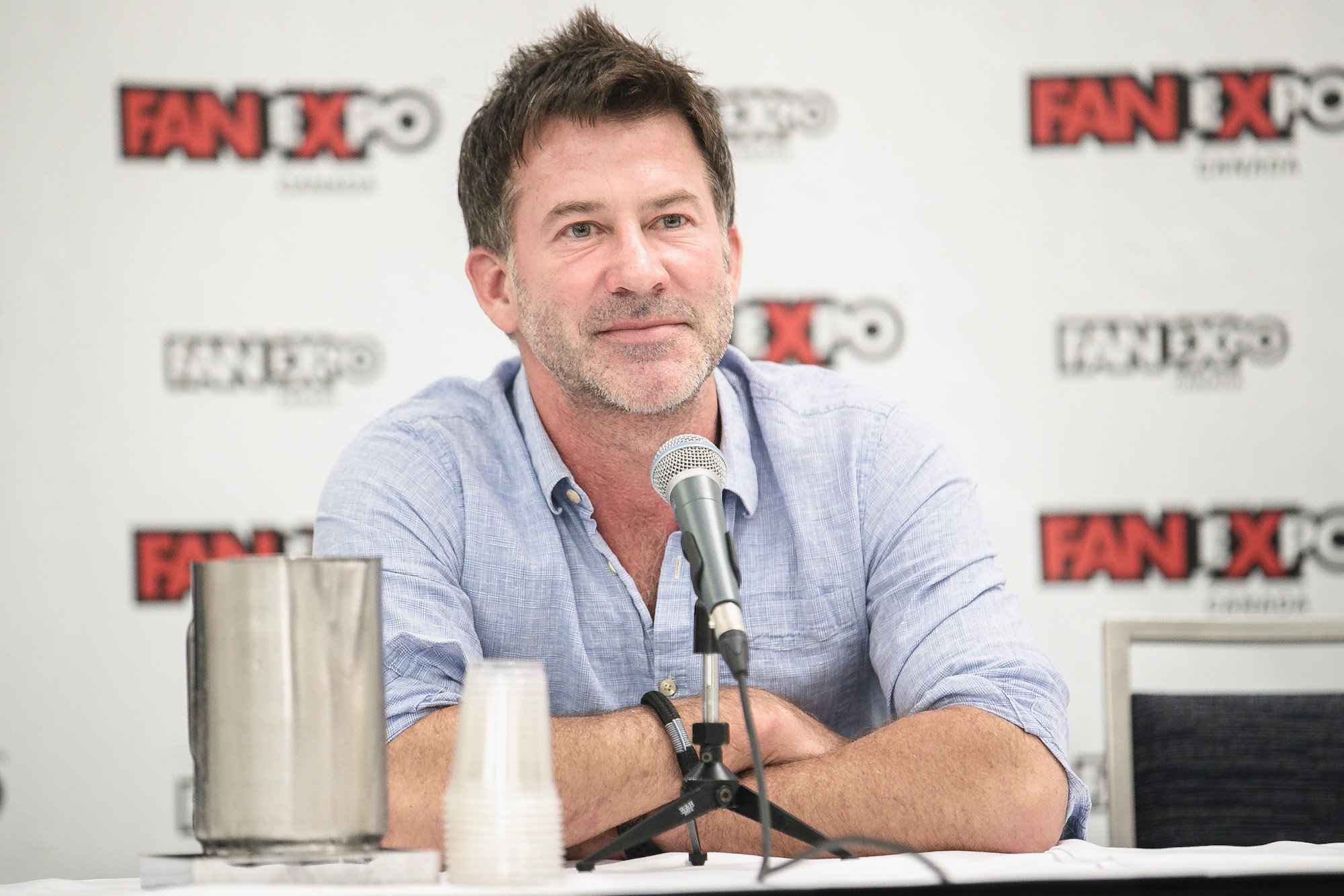 Joe Flanigan smiling in front of a white background with repeating logos