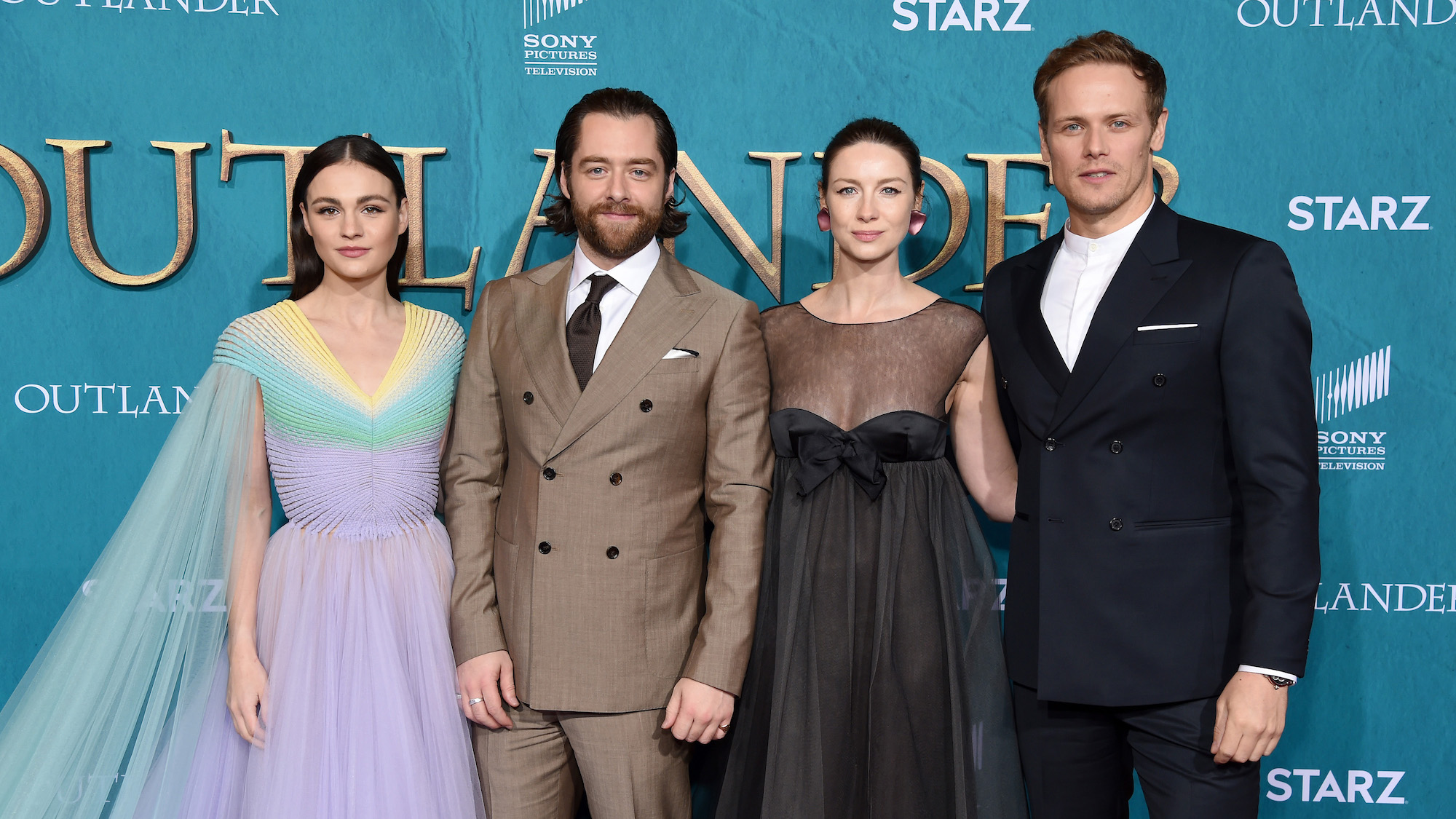 'Outlander' stars Sophie Skelton, Richard Rankin, Caitriona Balfe, and Sam Heughan smiling in front of a blue background in 2020
