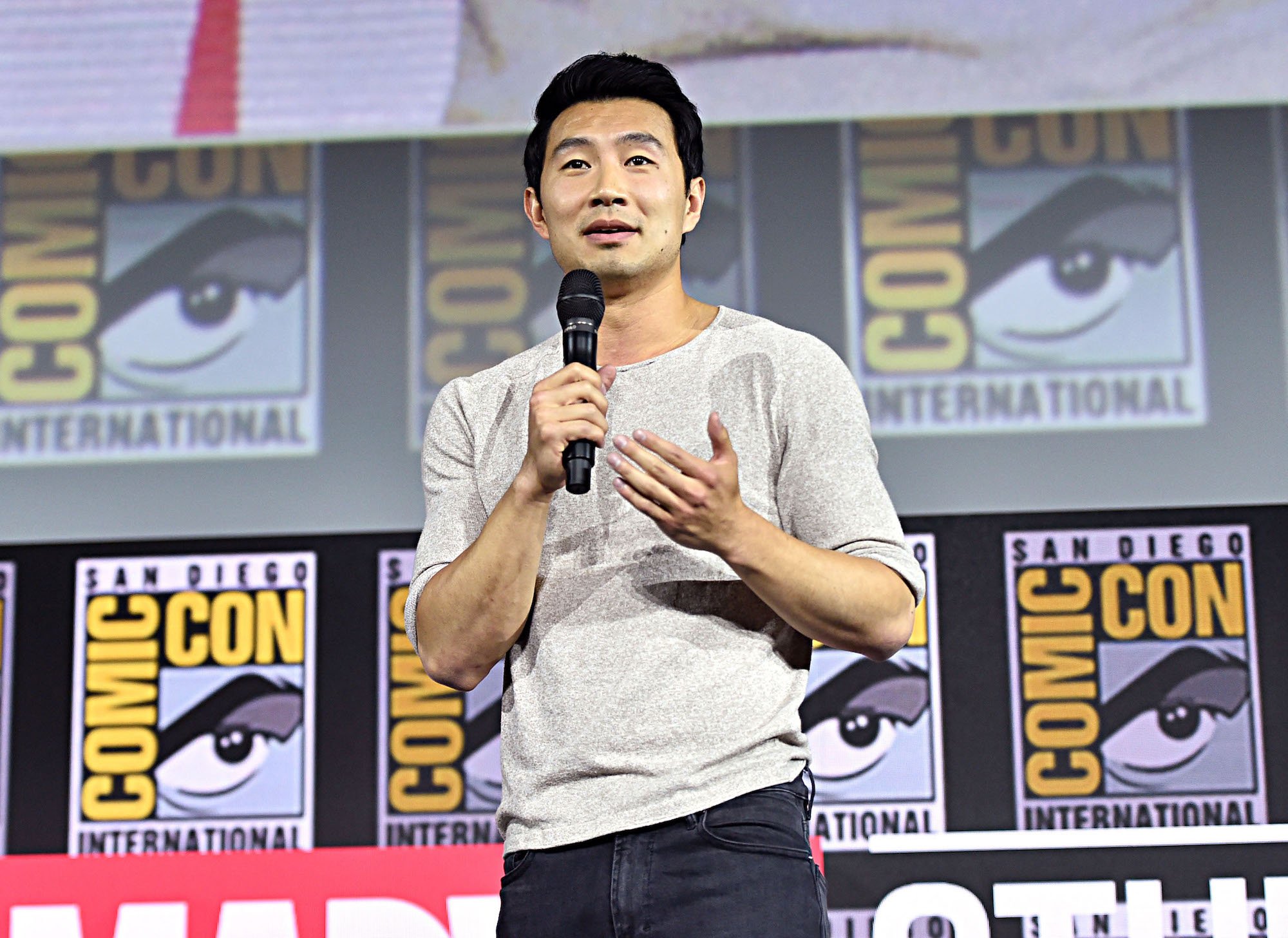 Simu Liu in front of a Comic Con background, speaking into a microphone