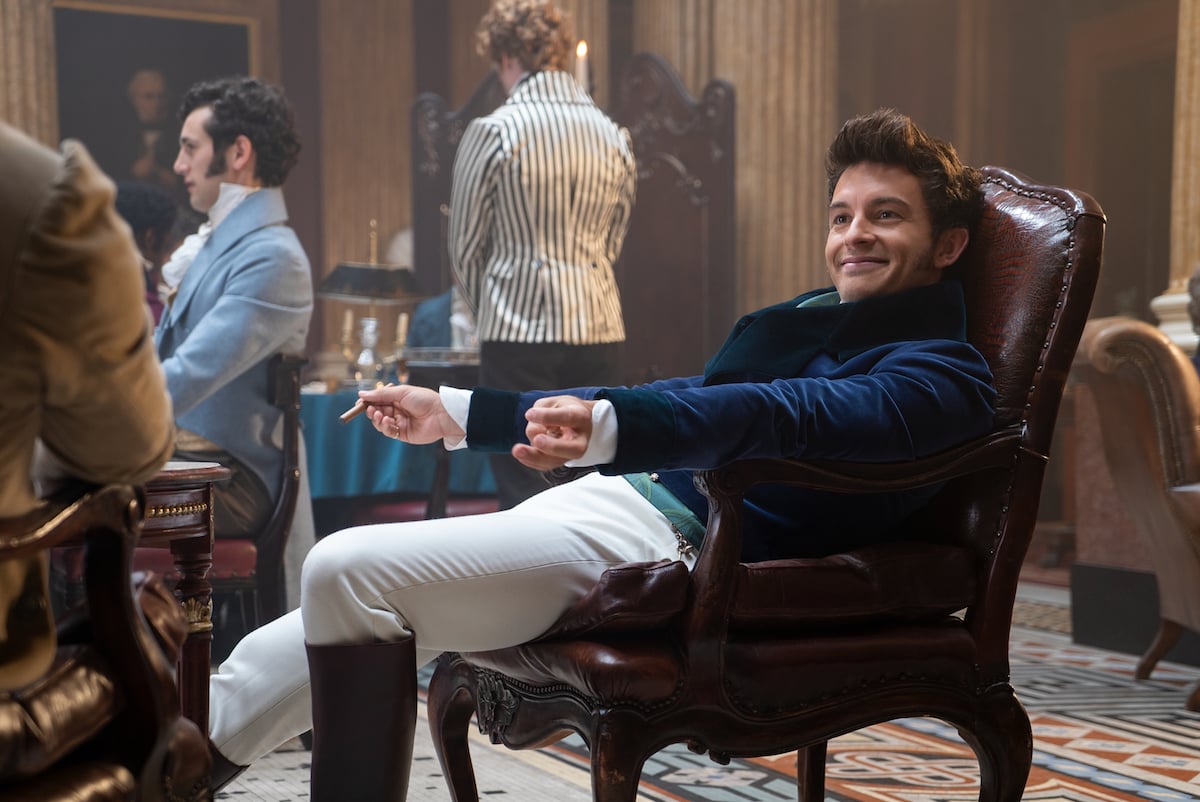 Smiling Anthony Bridgerton (Jonathan Bailey) wearing white pants and blue jacket sitting in a chair.