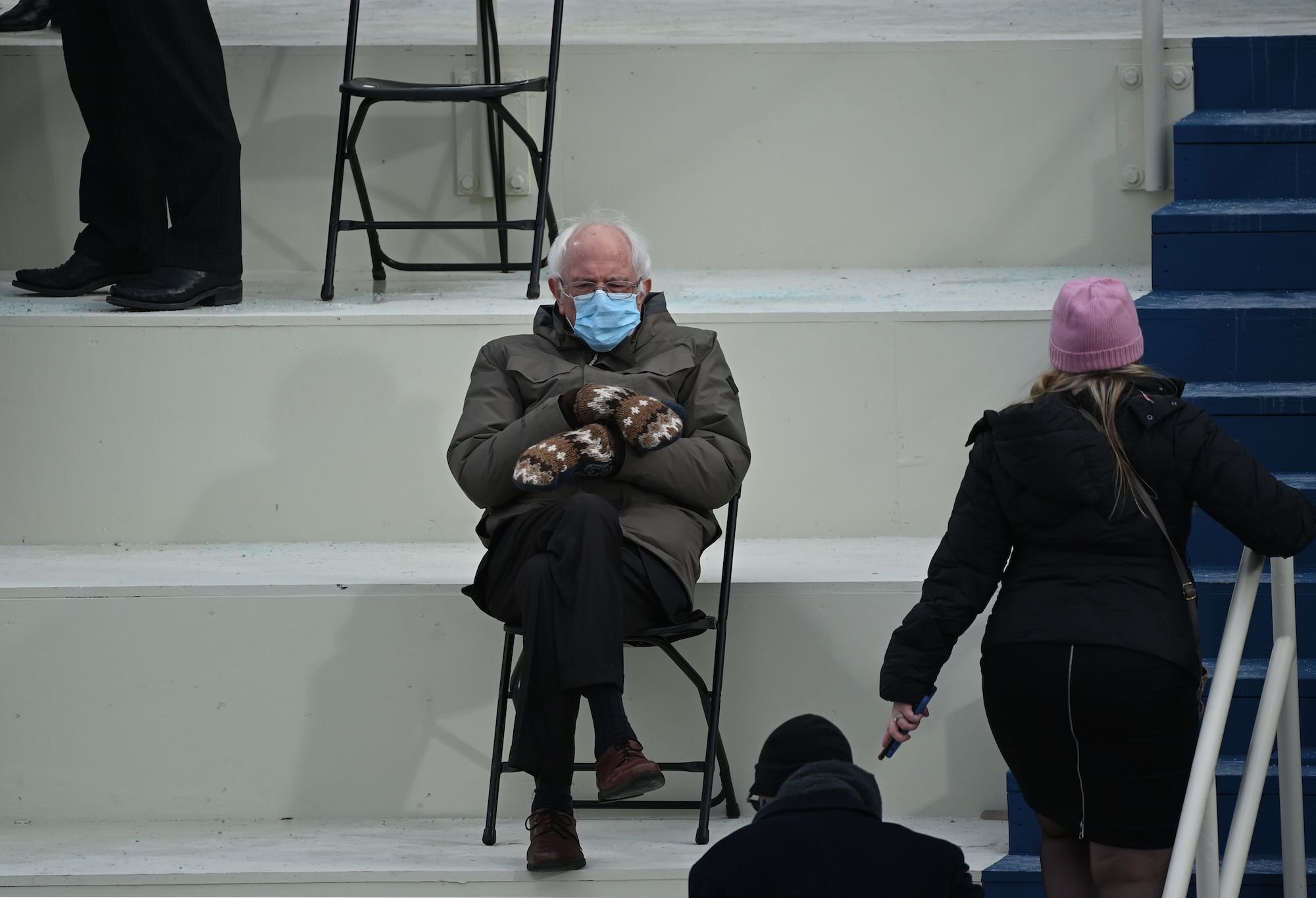 Senator Bernie Sanders sitting in a folding chair with arms crossed, wearing mittens, wearing a surgical mask