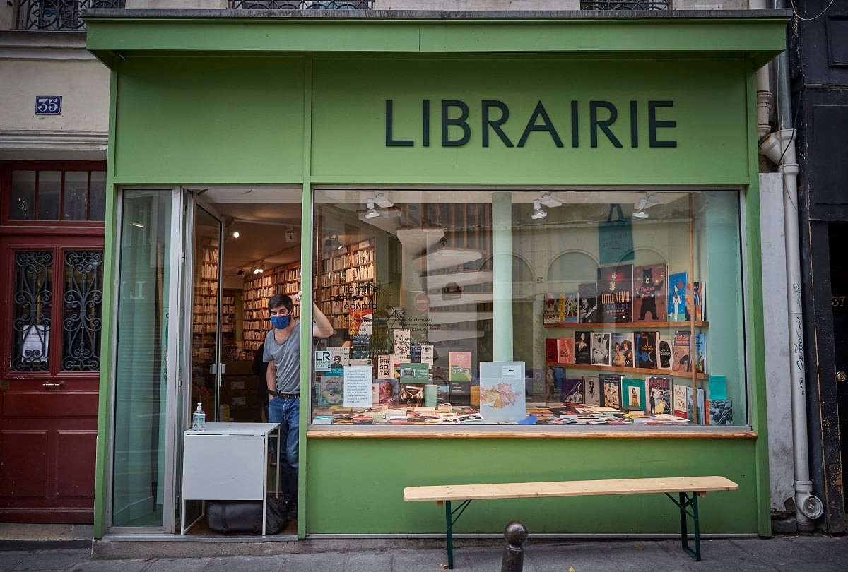 The street view of a glass-windowed bookshop in Paris named 'Librarie.' The storefront is green. A customer in a mask is inside the store.