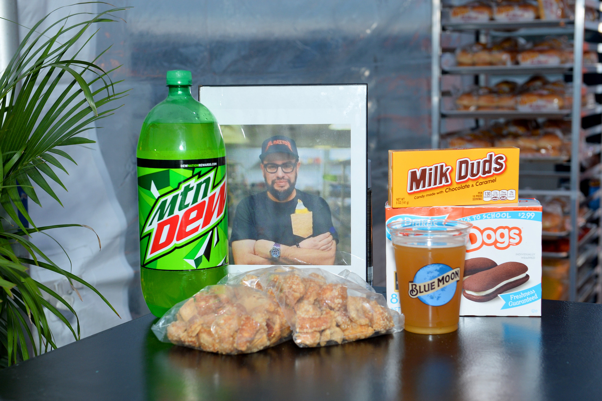 A memorial for Chef Carl Ruiz featuring a framed photo of him surrounded by a bottle of Mountain Dew, a cup of beer, a box of Milk Duds and a box of Devil Dogs