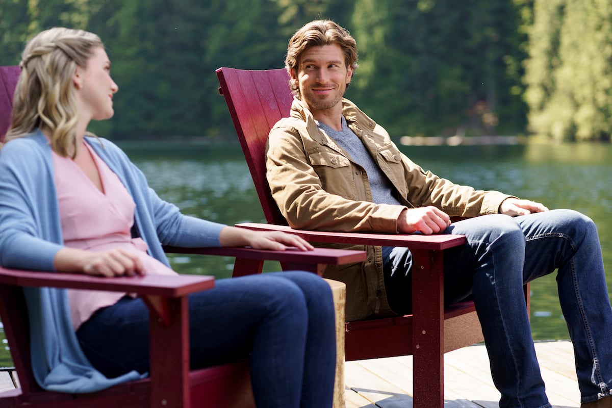 Man and woman sitting in red Adirondack chairs on a dock with lack in background