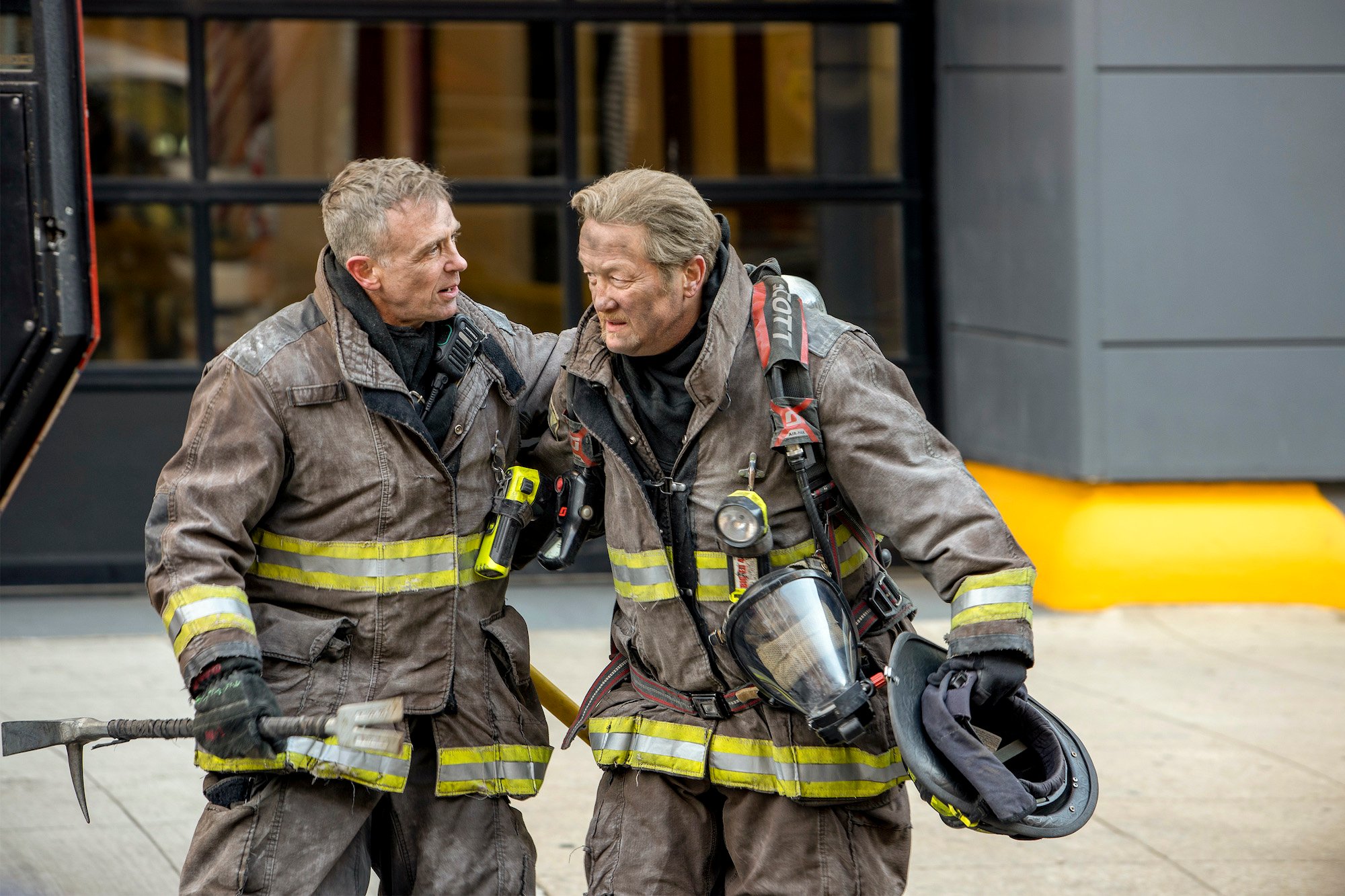 (L-R) David Eigenberg as Christopher Herrmann, Christian Stolte as Randall “Mouch” McHolland in full firefighter gear, covered in soot, standing on a sidewalk talking