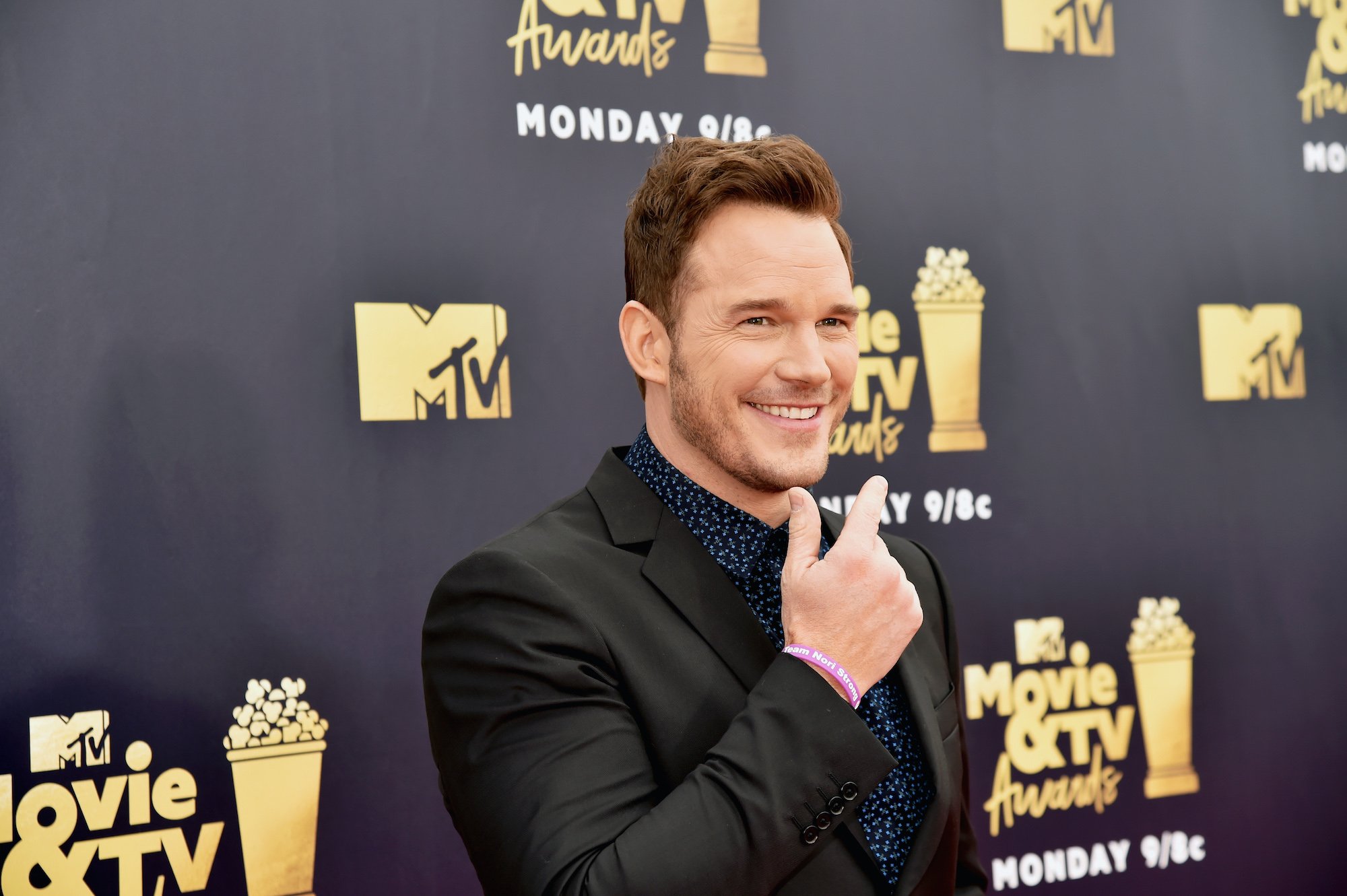Chris Pratt smiling in front of a dark gray background with repeating gold logos